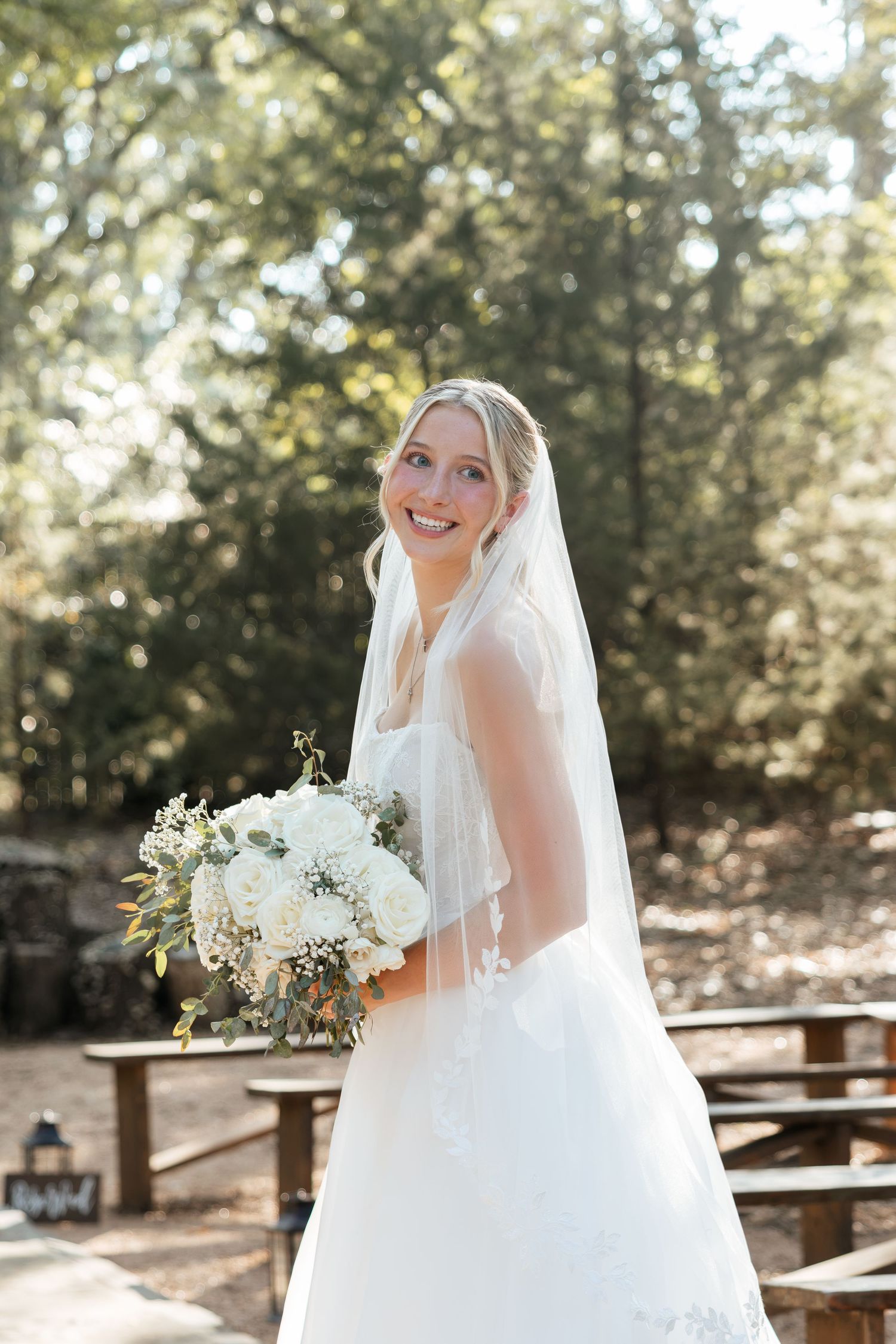 Smiling bride in white wedding gown and veil holding white floral bouquet outdoors in sunlit forest setting.