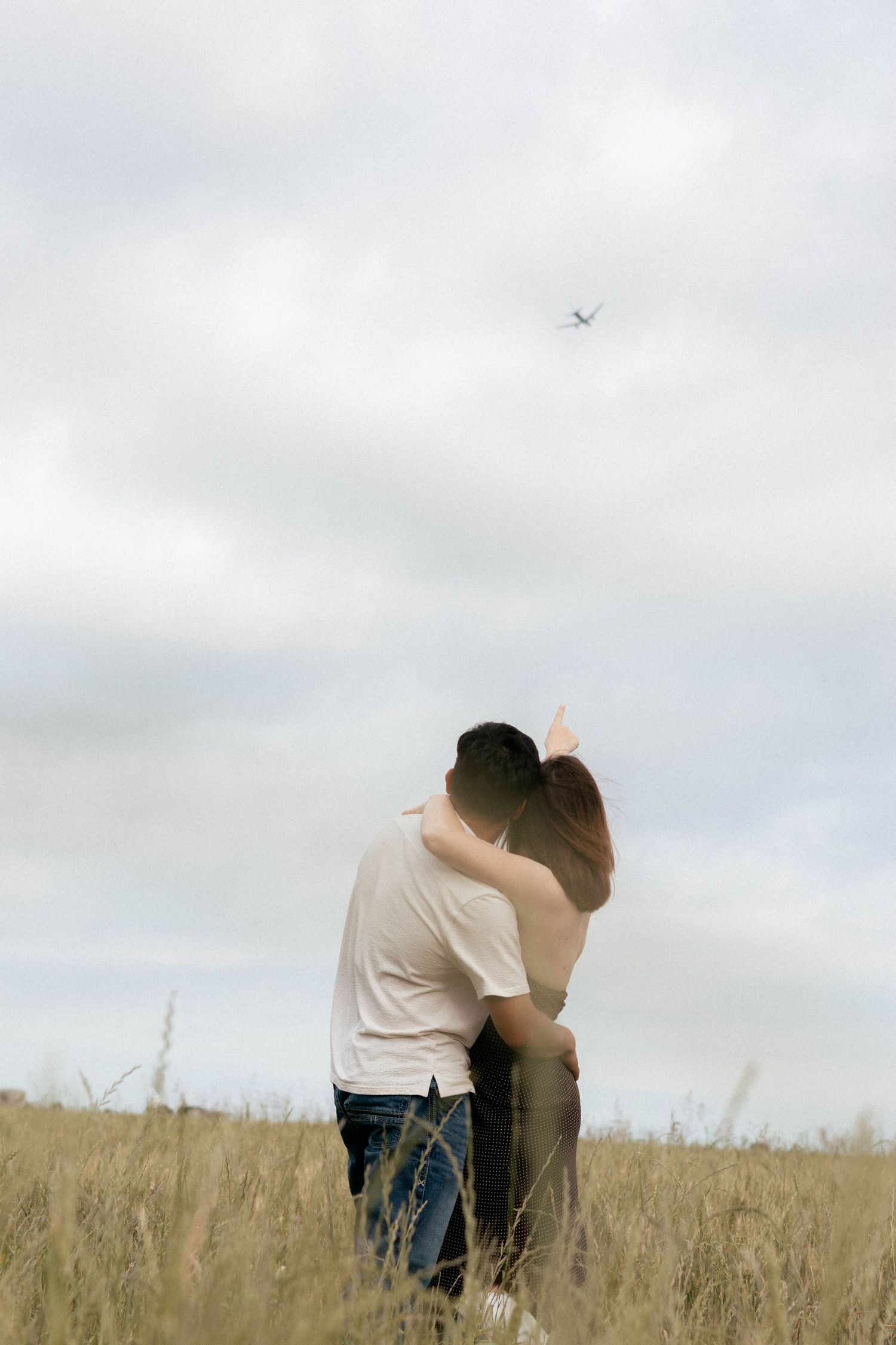 Couple embracing romantically in a golden field under cloudy skies with an airplane flying overhead.