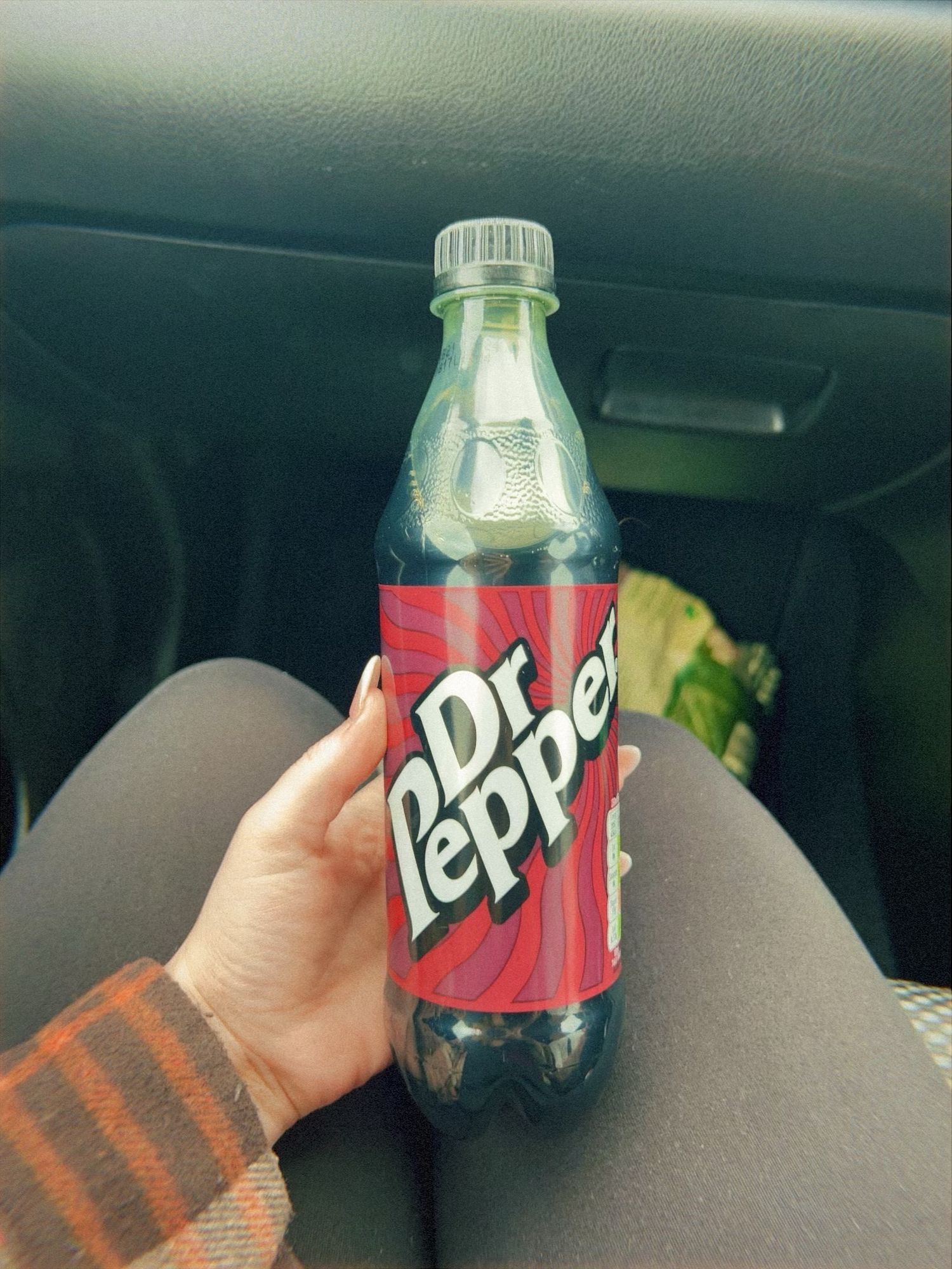 Hand holding a bottle of Dr Pepper soda against a car interior background with gray upholstery.