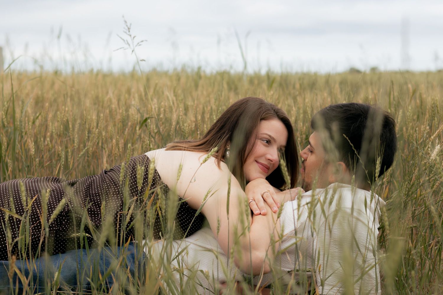 Couple lying in a golden wheat field, holding hands and gazing at each other romantically on a cloudy day.
