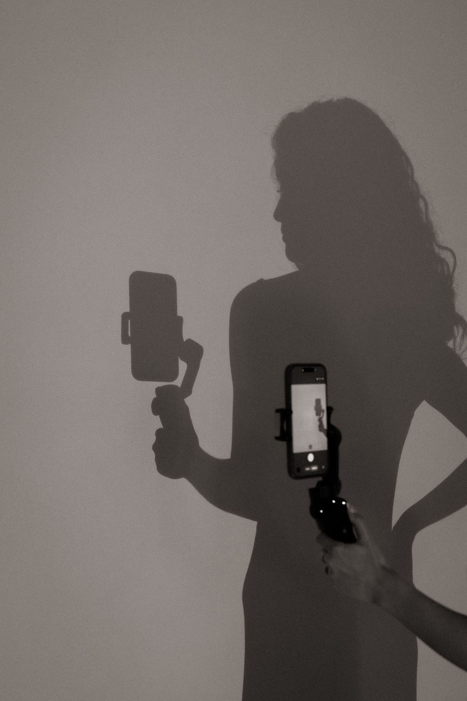Artistic silhouette of a woman holding a smartphone gimbal stabilizer against a neutral wall.