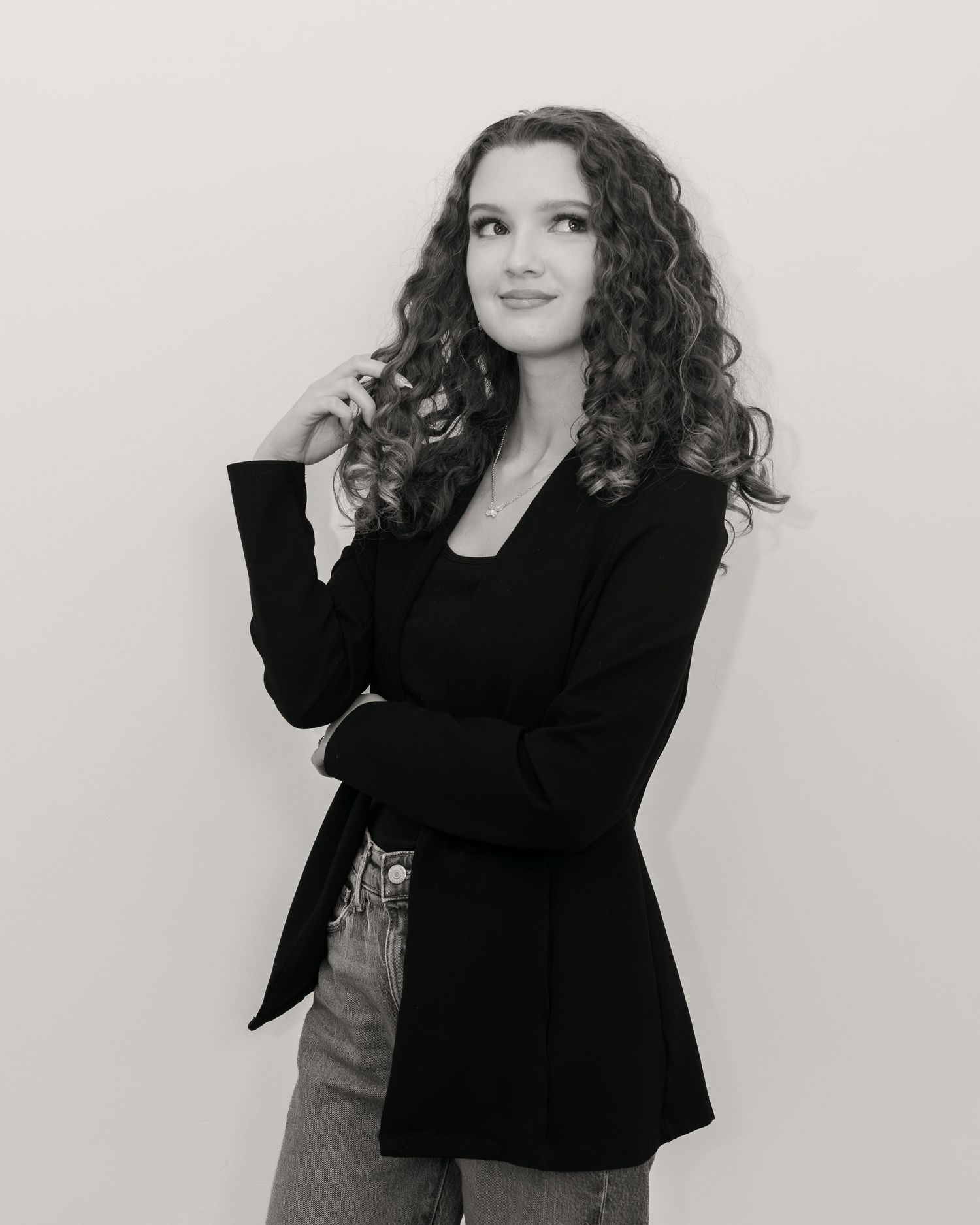 Black and white portrait of curly-haired young woman in black blazer posing thoughtfully against white background.