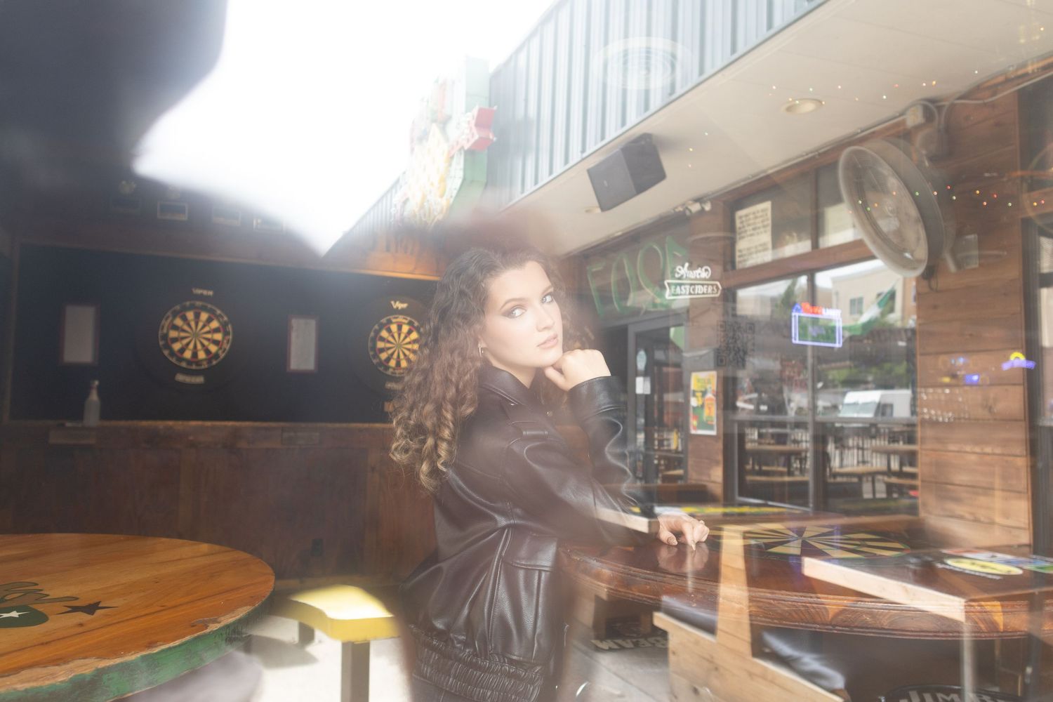 Young woman in black leather jacket poses inside a bar with dartboard and wooden furniture visible.