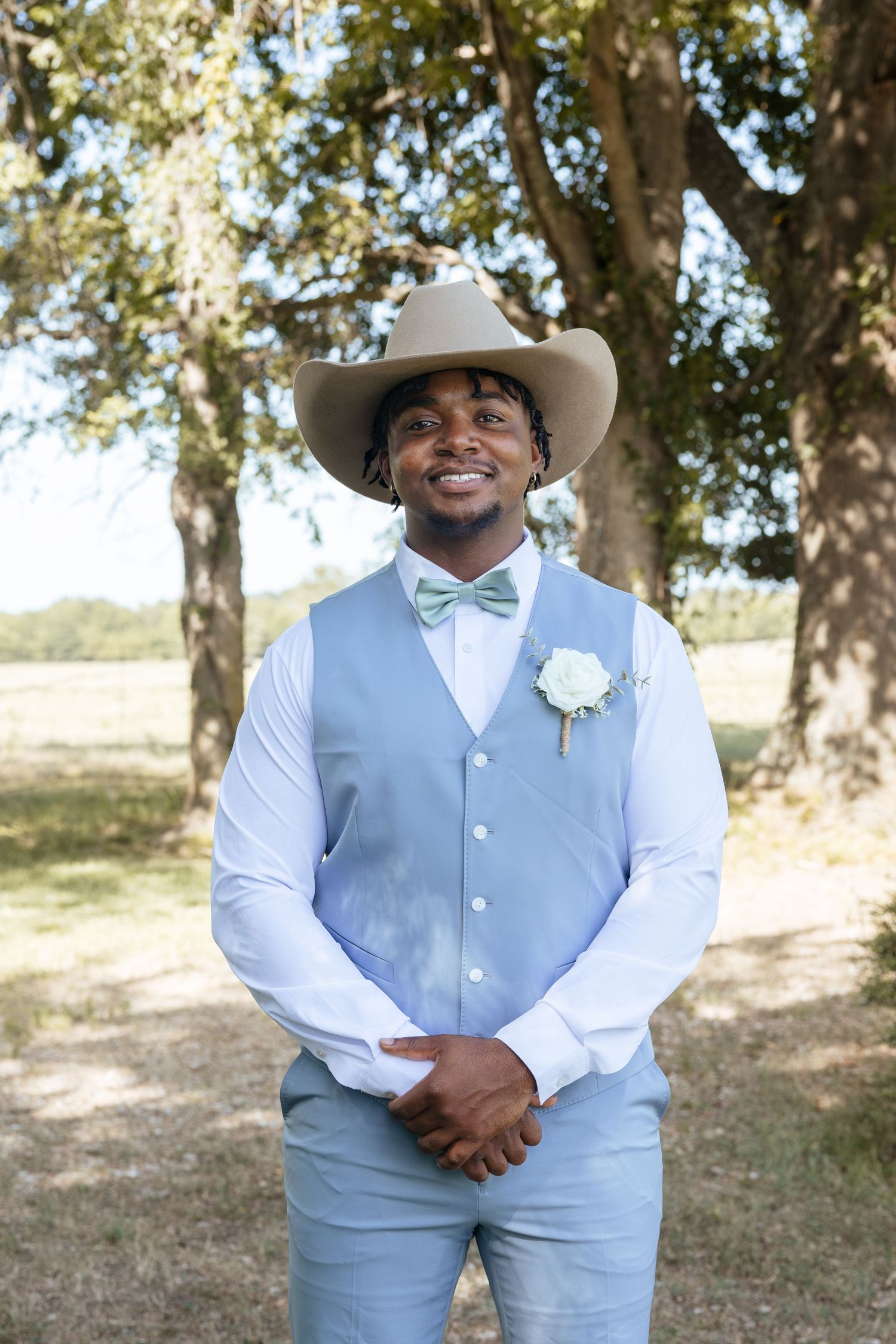 Smiling groom in light blue vest, bow tie, and cowboy hat with boutonniere, standing outdoors under trees.
