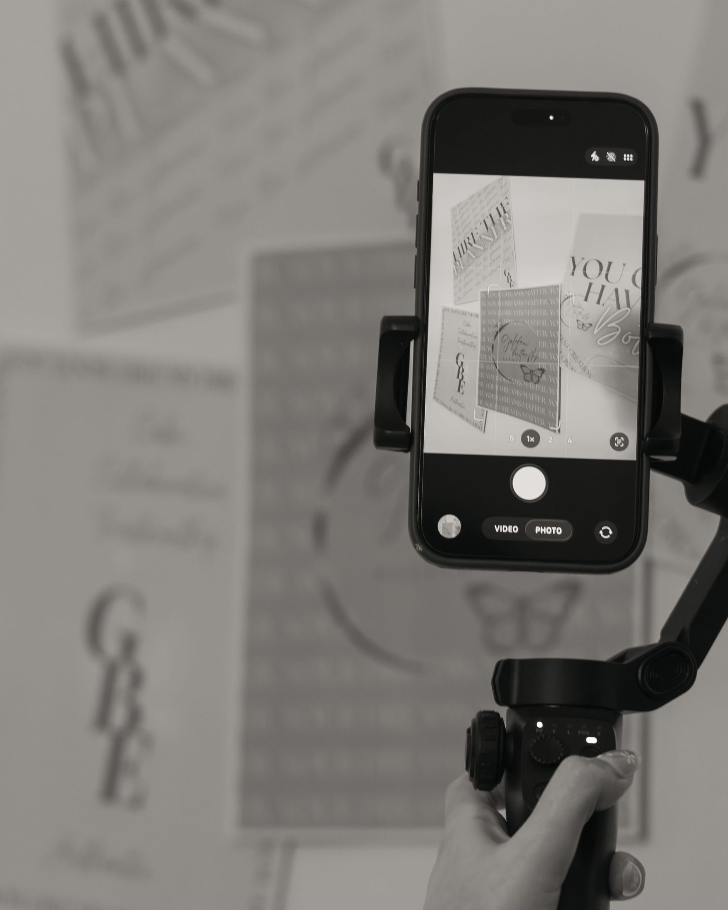A smartphone mounted on a gimbal stabilizer photographs decorative text posters in a black and white studio setting.