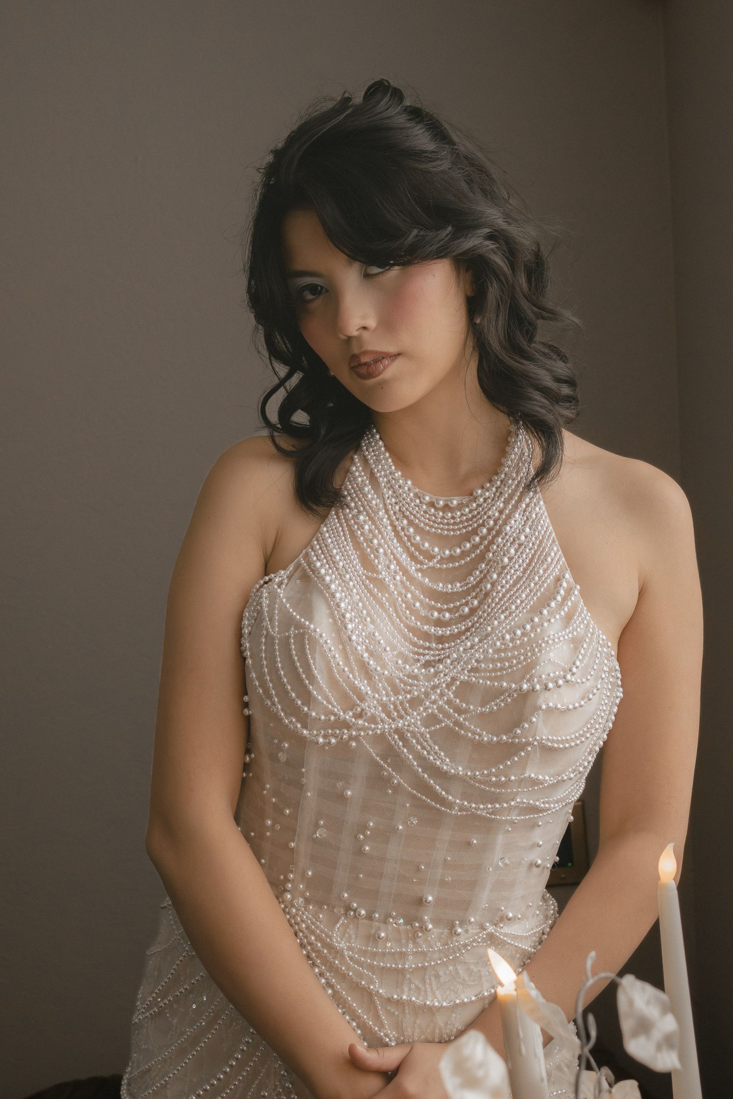 Bride in elegant pearl-draped halter wedding gown holding a candle in a softly lit studio setting.