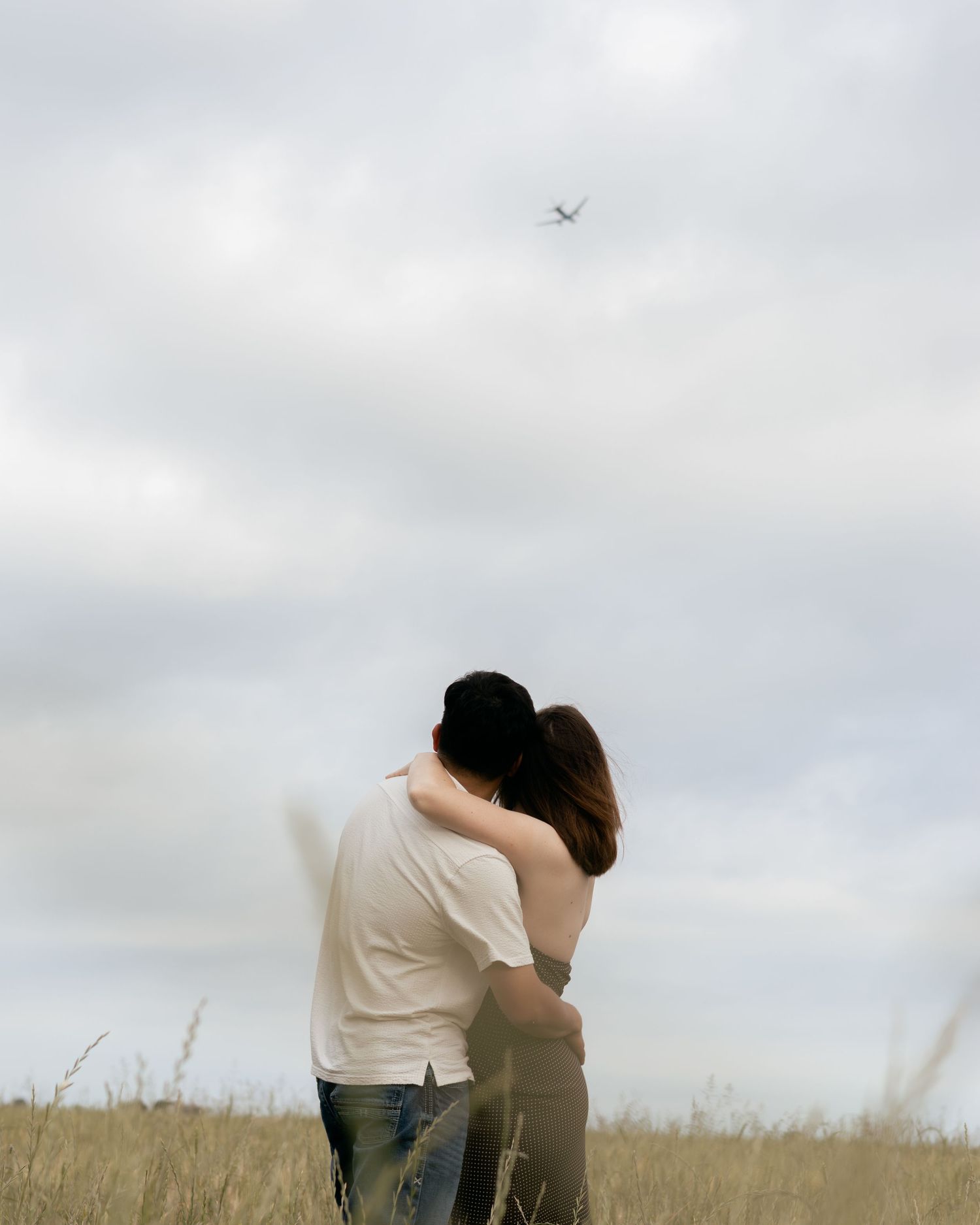 Couple embracing in a golden field under cloudy skies with a bird flying overhead.