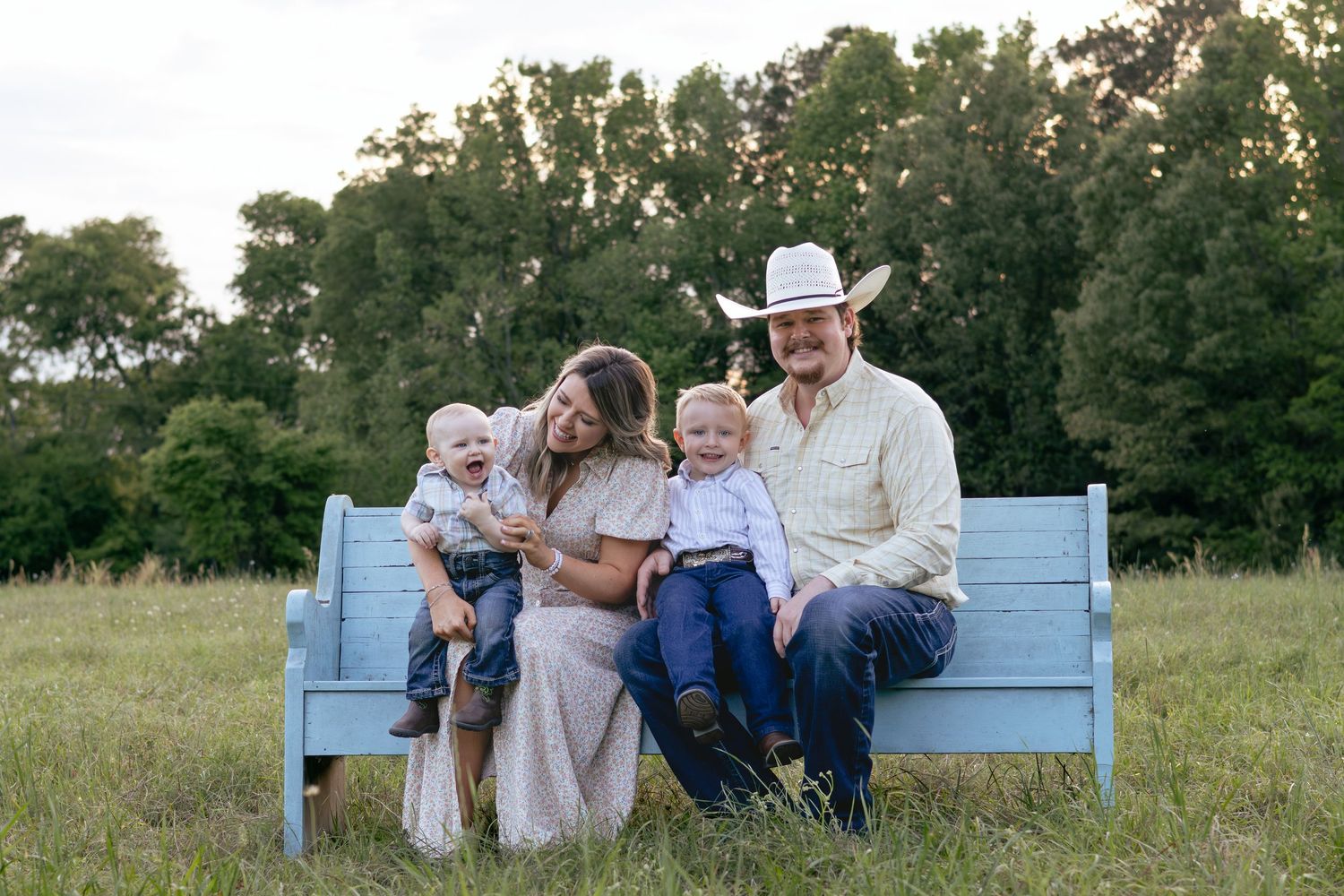 Family of four sitting on a blue bench in a green field, parents with two young children in rustic country attire.