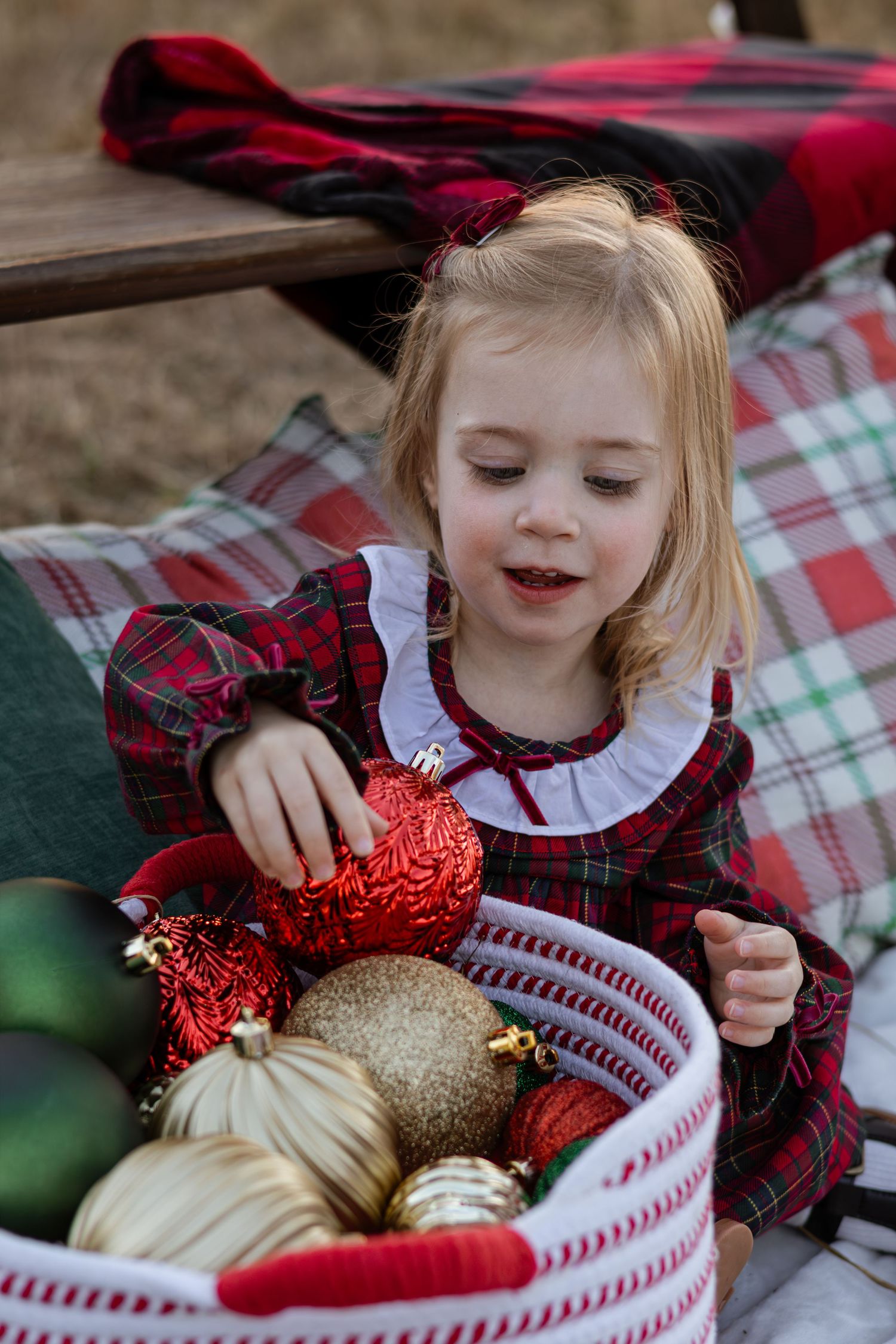 A small child in a red plaid dress examines holiday ornaments in a white basket with festive decorations.