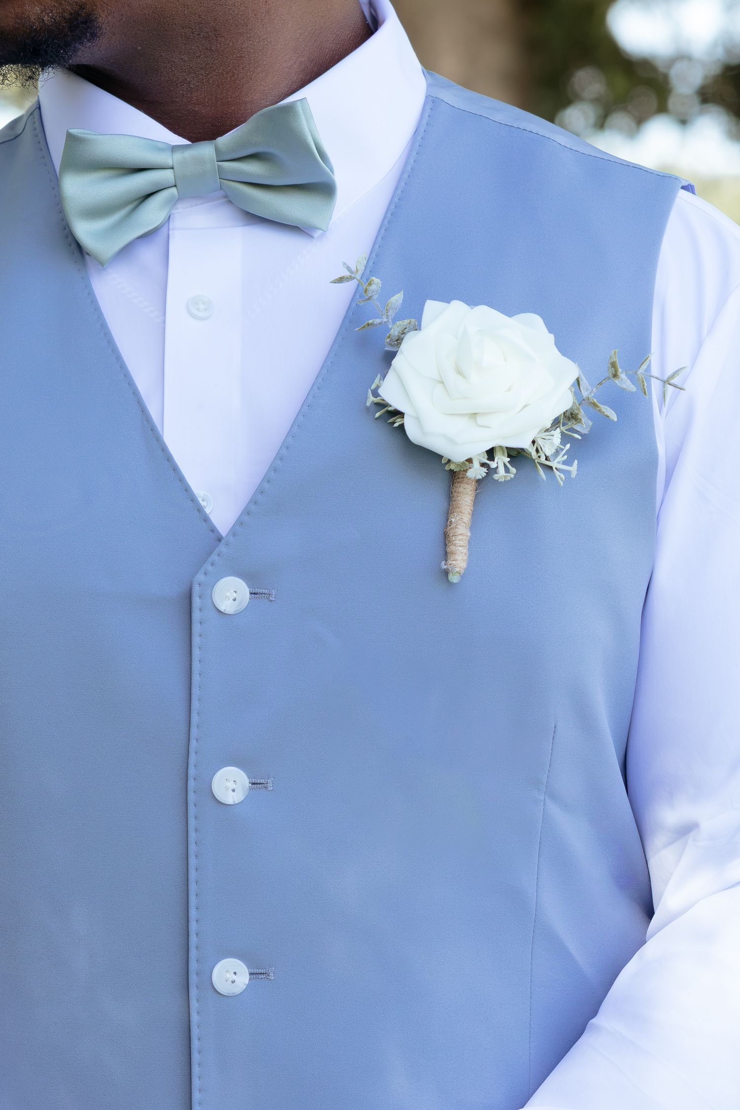 Groom wearing light blue vest, sage green bow tie, and white rose boutonniere at wedding.