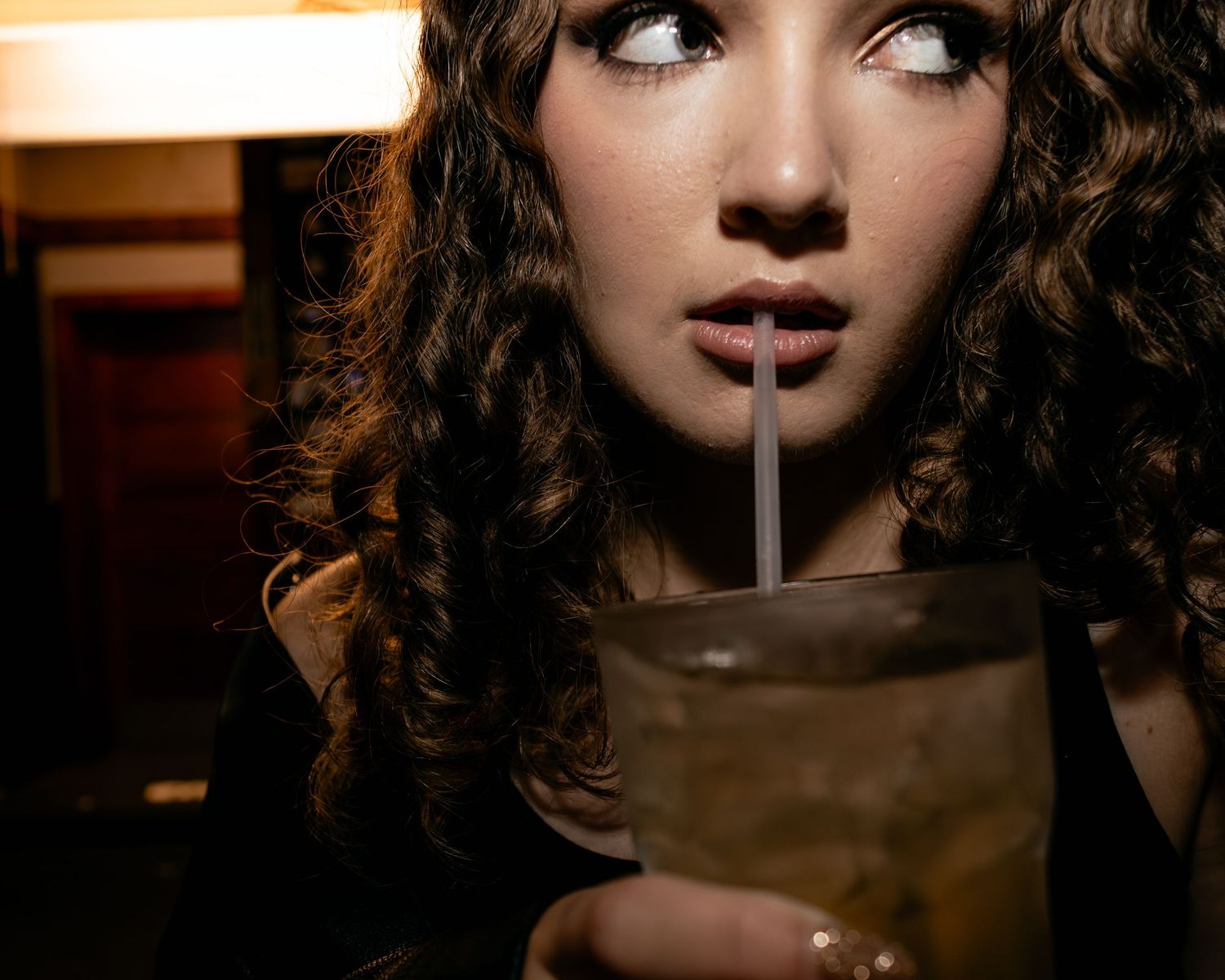 Young woman with curly hair sipping a drink through a straw at a dimly lit bar.