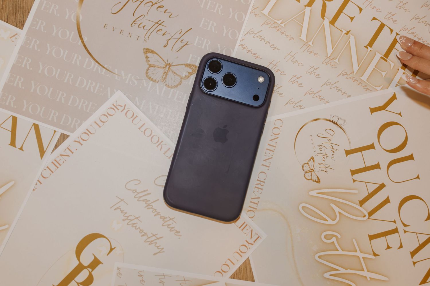 A dark iPhone with triple camera sits atop scattered gold foil motivational quote cards on a white surface.