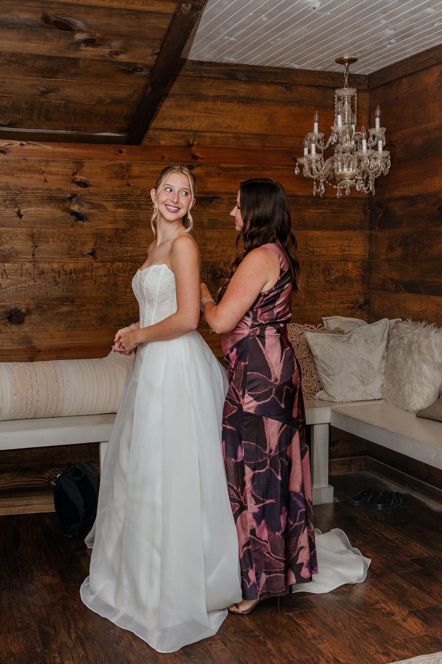 Bride in white strapless gown being helped by bridesmaid in floral dress inside rustic wood-paneled bridal suite.