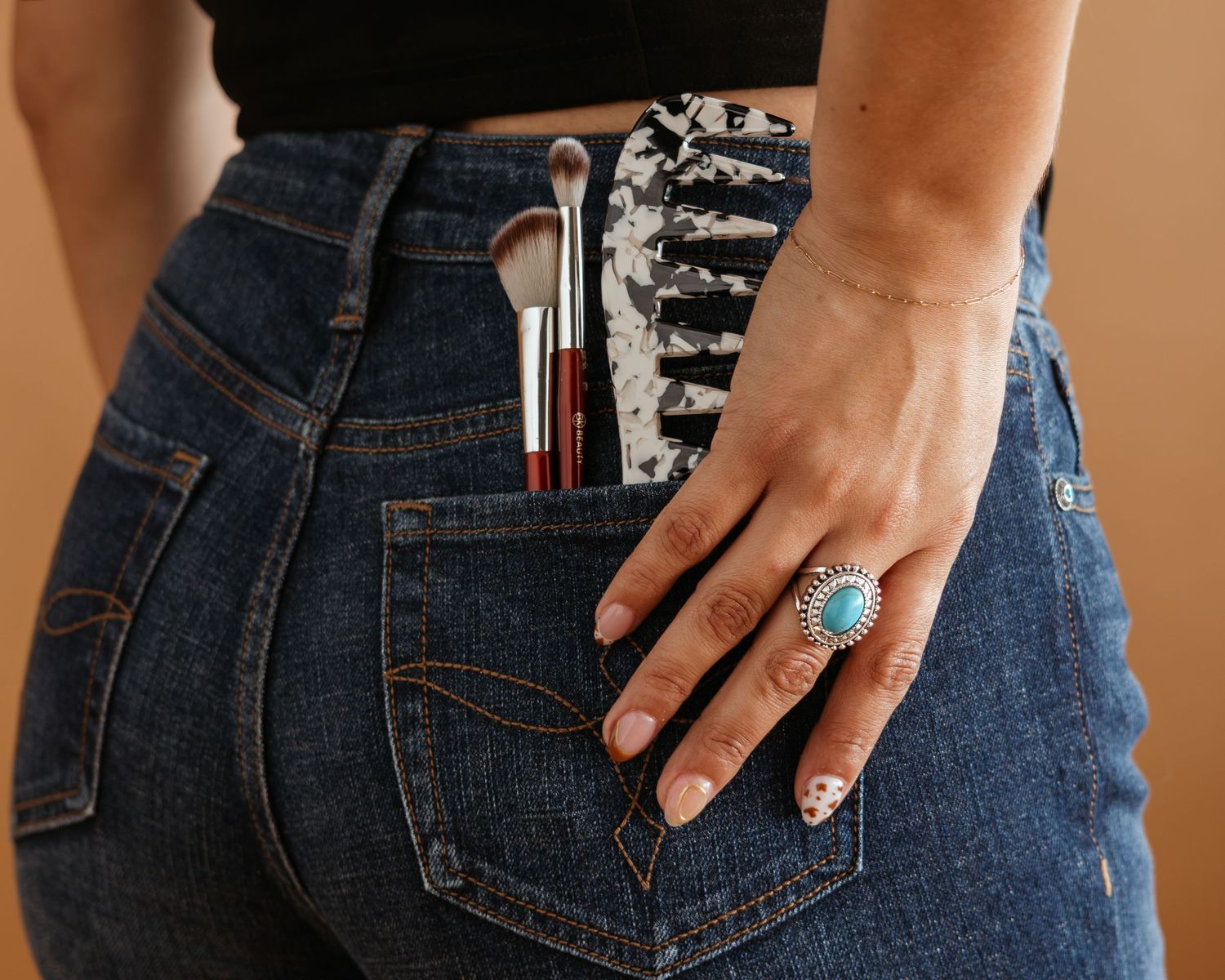 Woman wearing jeans with makeup brushes in back pocket and turquoise silver ring on hand.