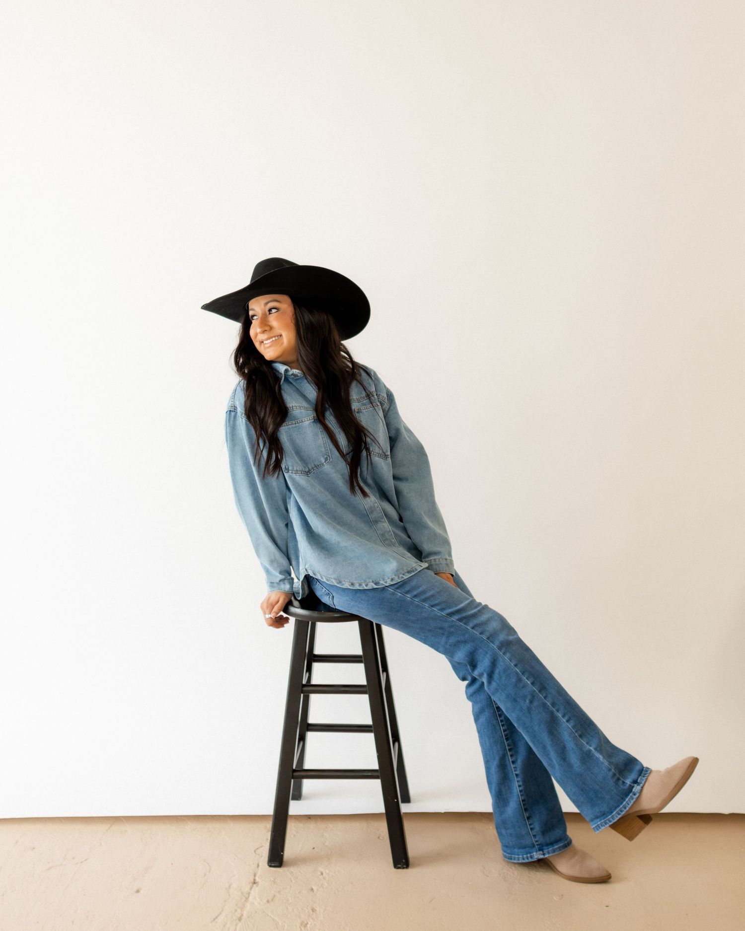 Woman in black wide-brim hat, denim shirt, flare jeans, and tan boots sitting on a black stool.