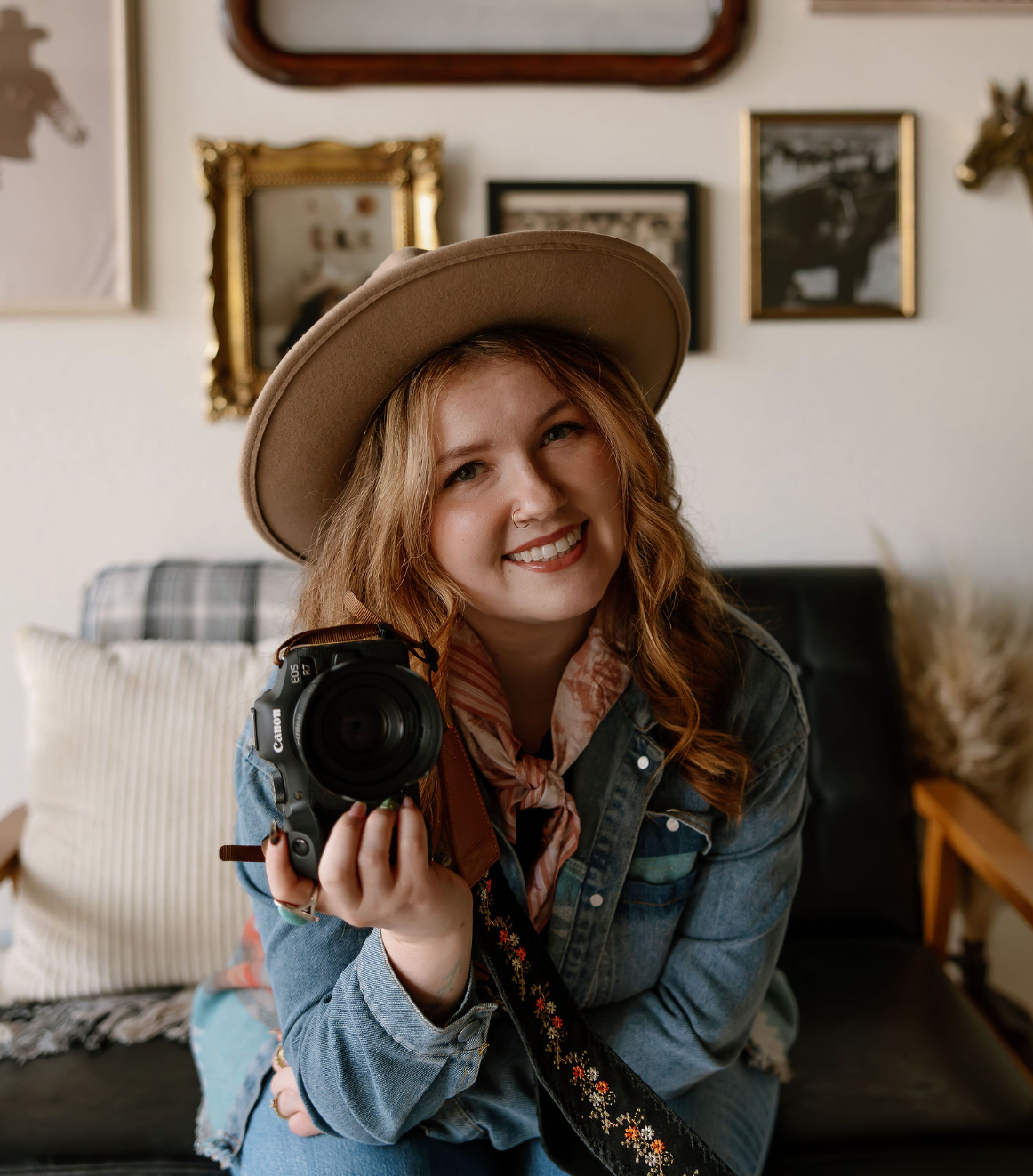A photographer wearing a denim jacket and brown hat laughs while holding a camera in an indoor setting.