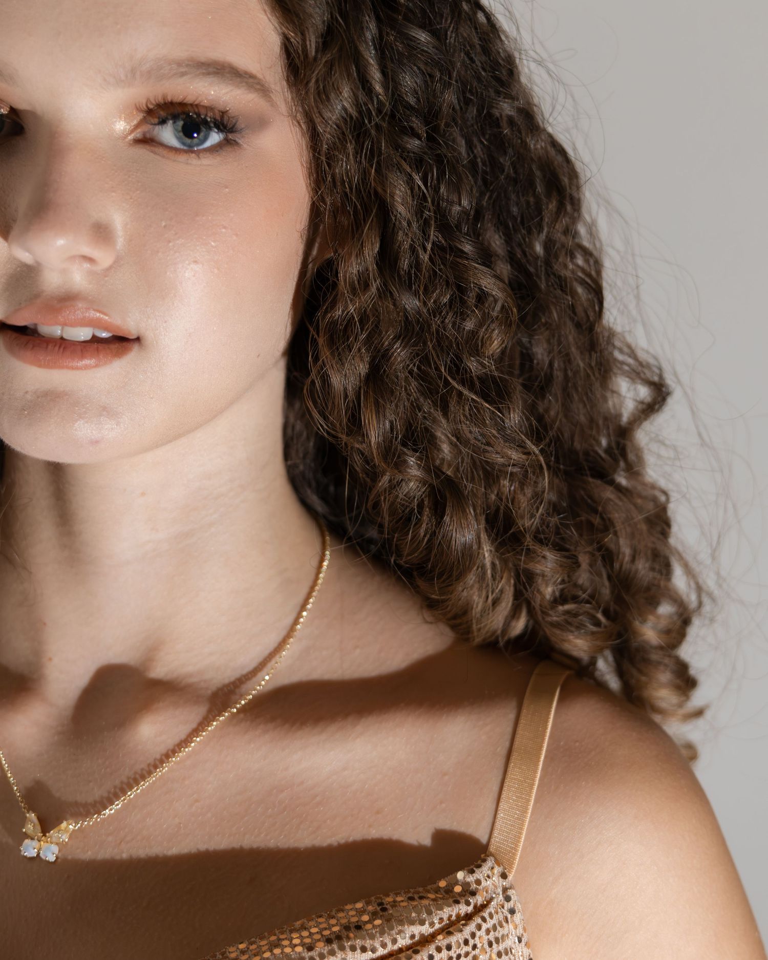 Young woman with curly brown hair wearing a gold sequin dress and delicate gold necklace poses for a close-up portrait.