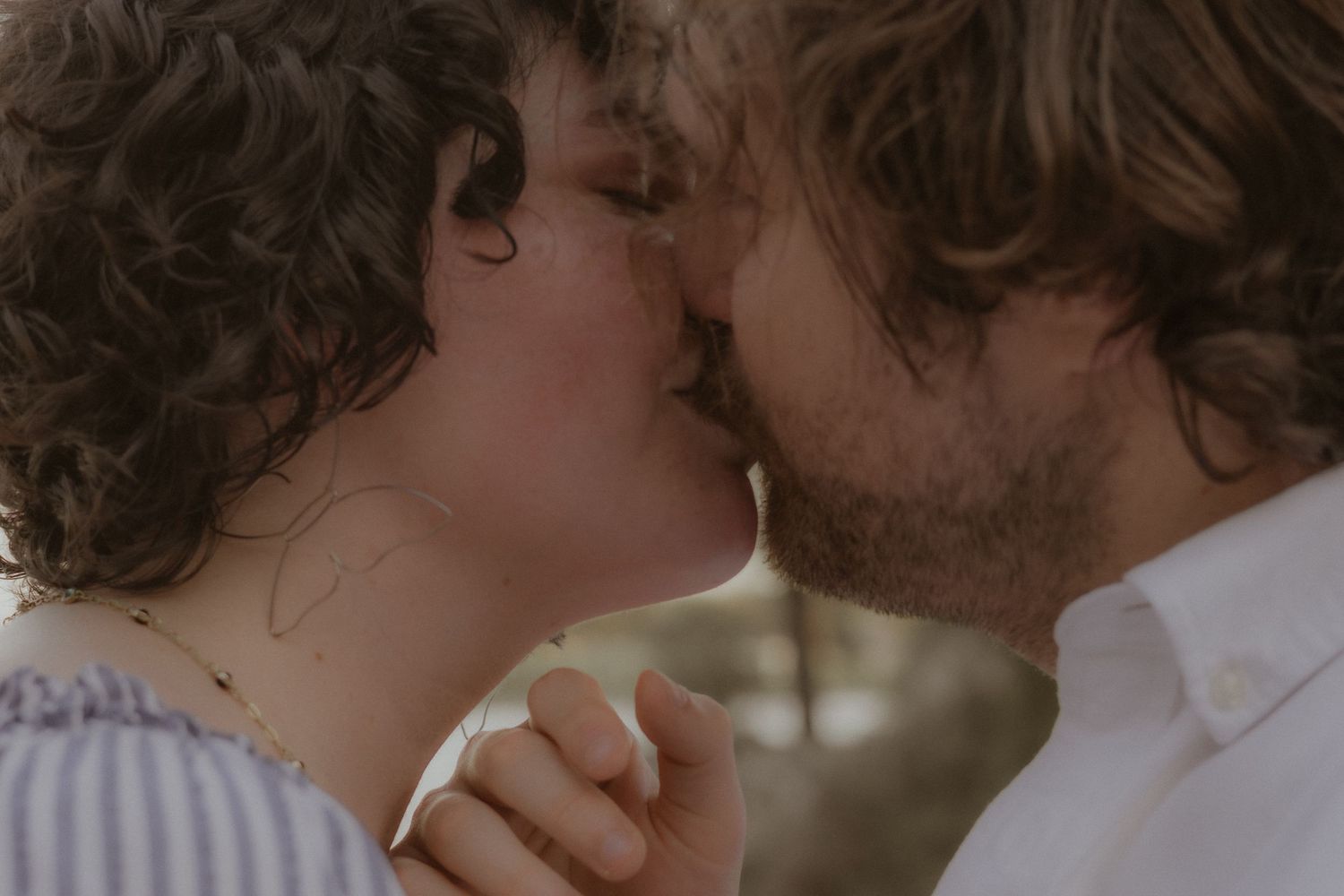 Romantic couple sharing a tender kiss outdoors, holding hands, with soft natural bokeh background.