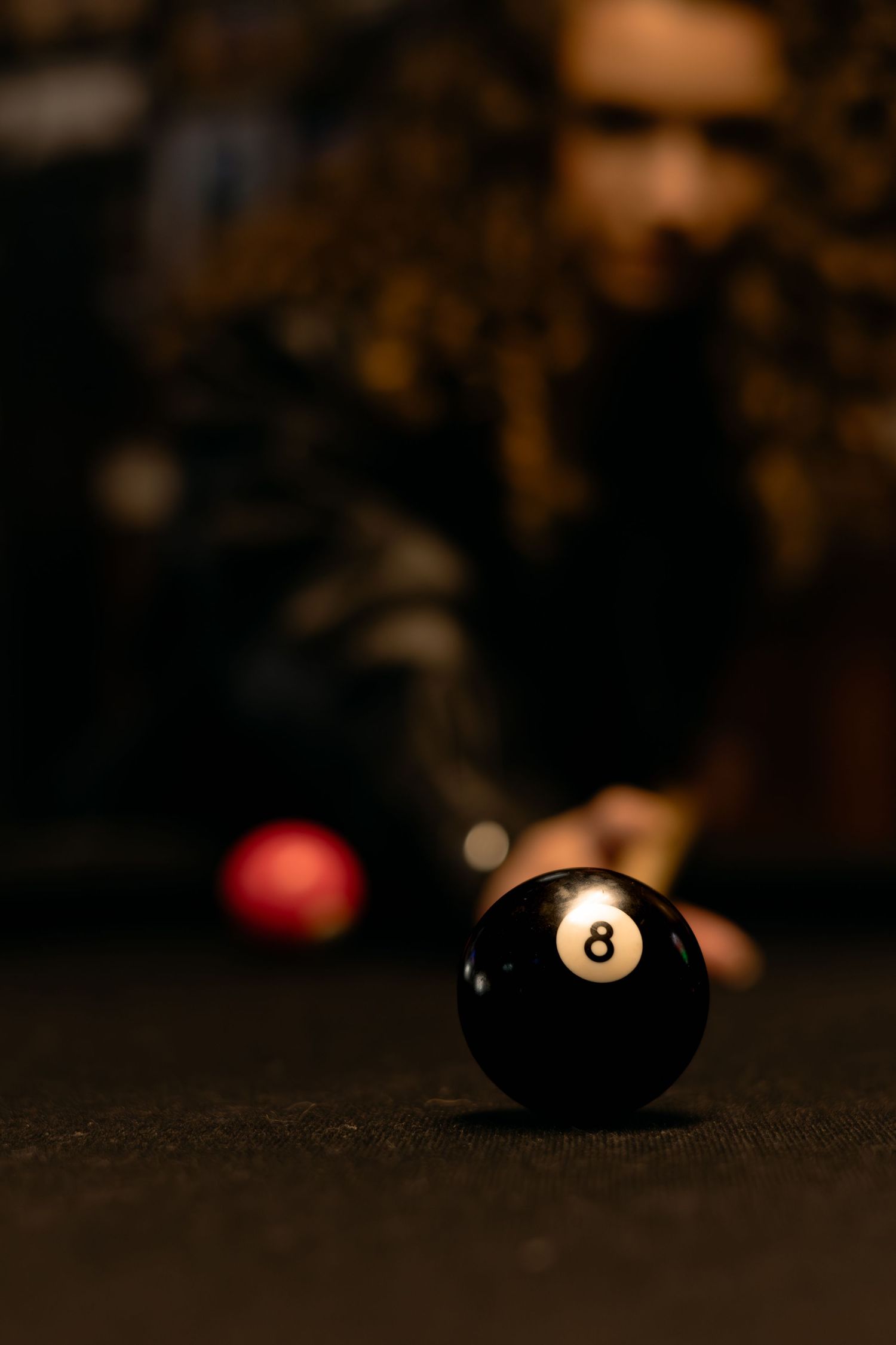 Close-up of an 8-ball on a pool table with a blurred player lining up a shot in the background.