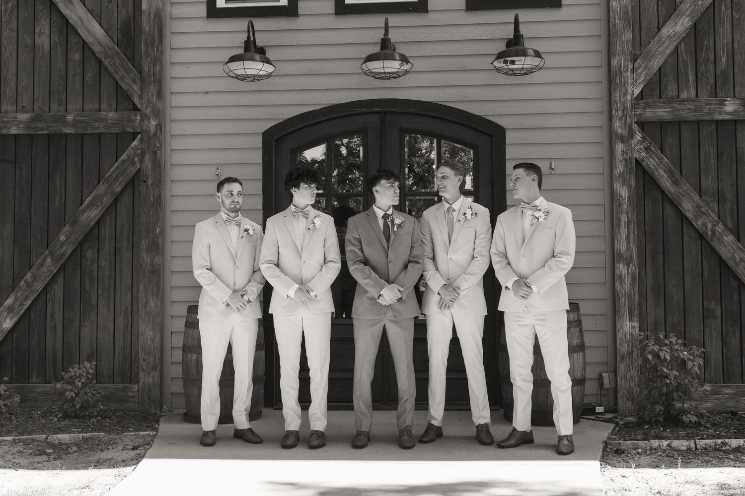 Five groomsmen in light suits pose in front of a rustic barn door in a classic black and white wedding photo.