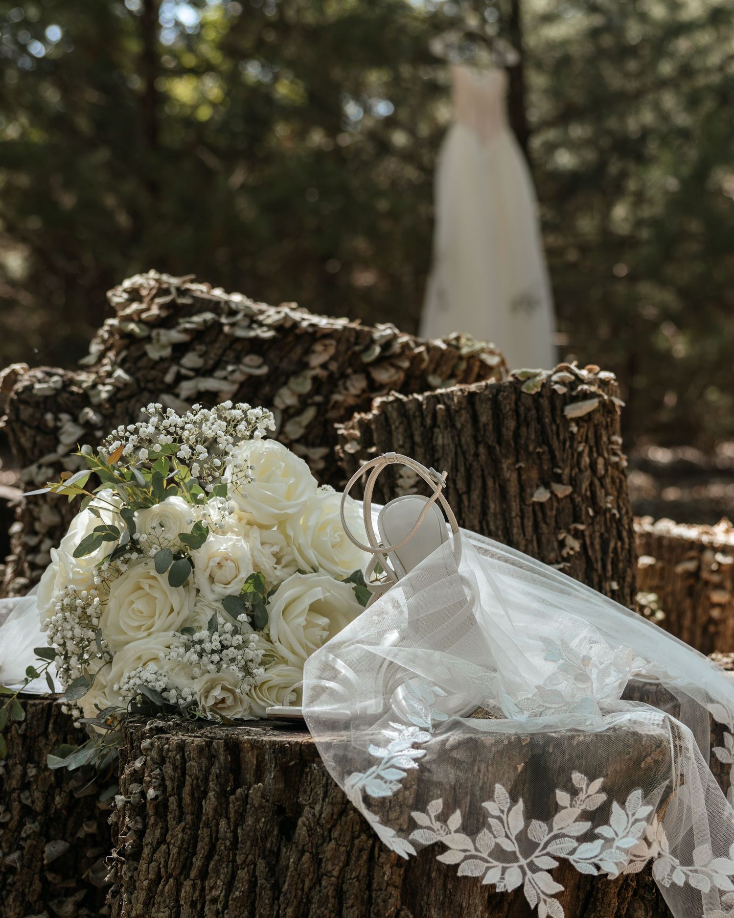 White rose bridal bouquet with wedding rings and veil on tree stump, wedding dress hanging in forest background.