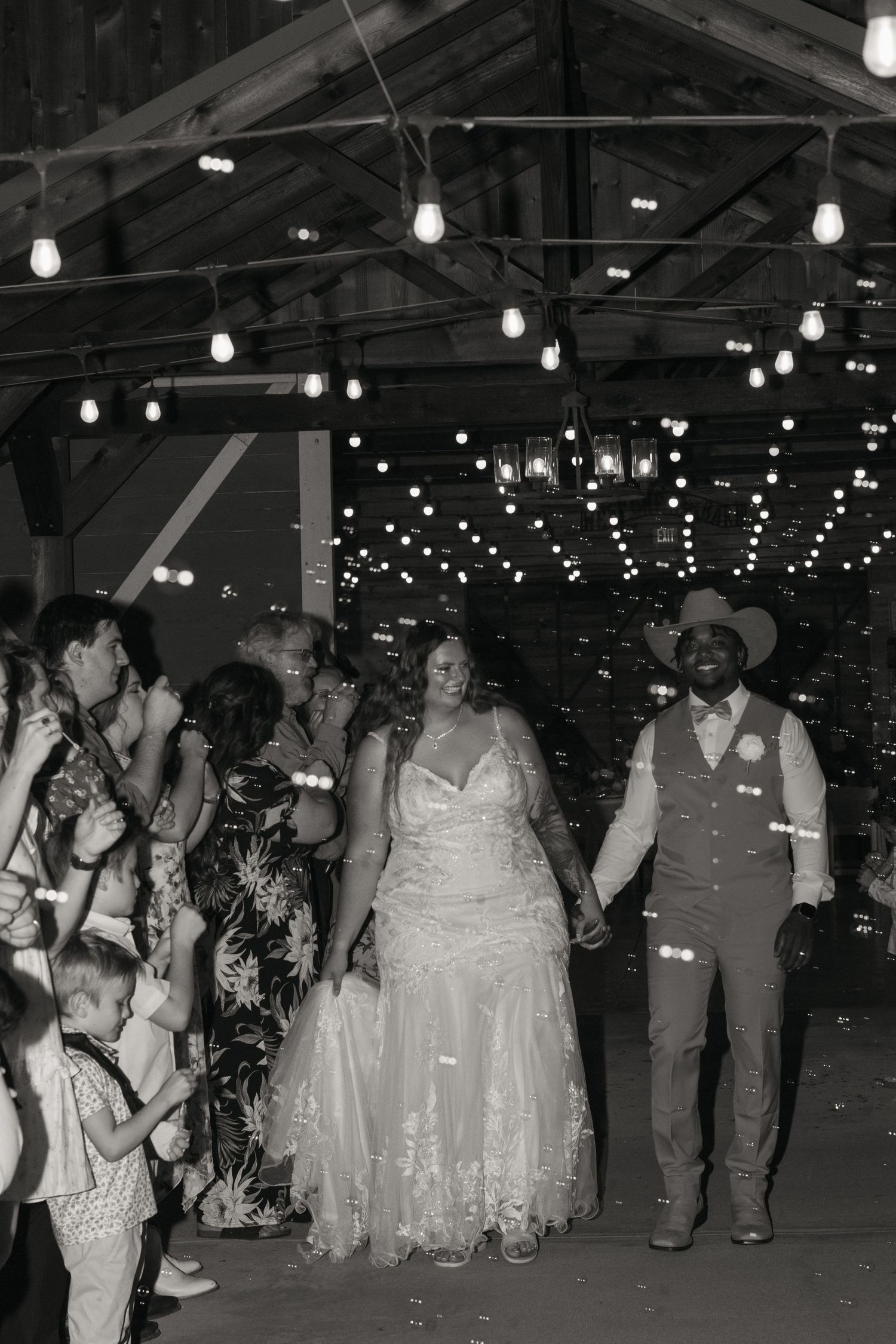 Bride and groom walking through bubble send-off in black and white photo inside warmly lit barn reception.