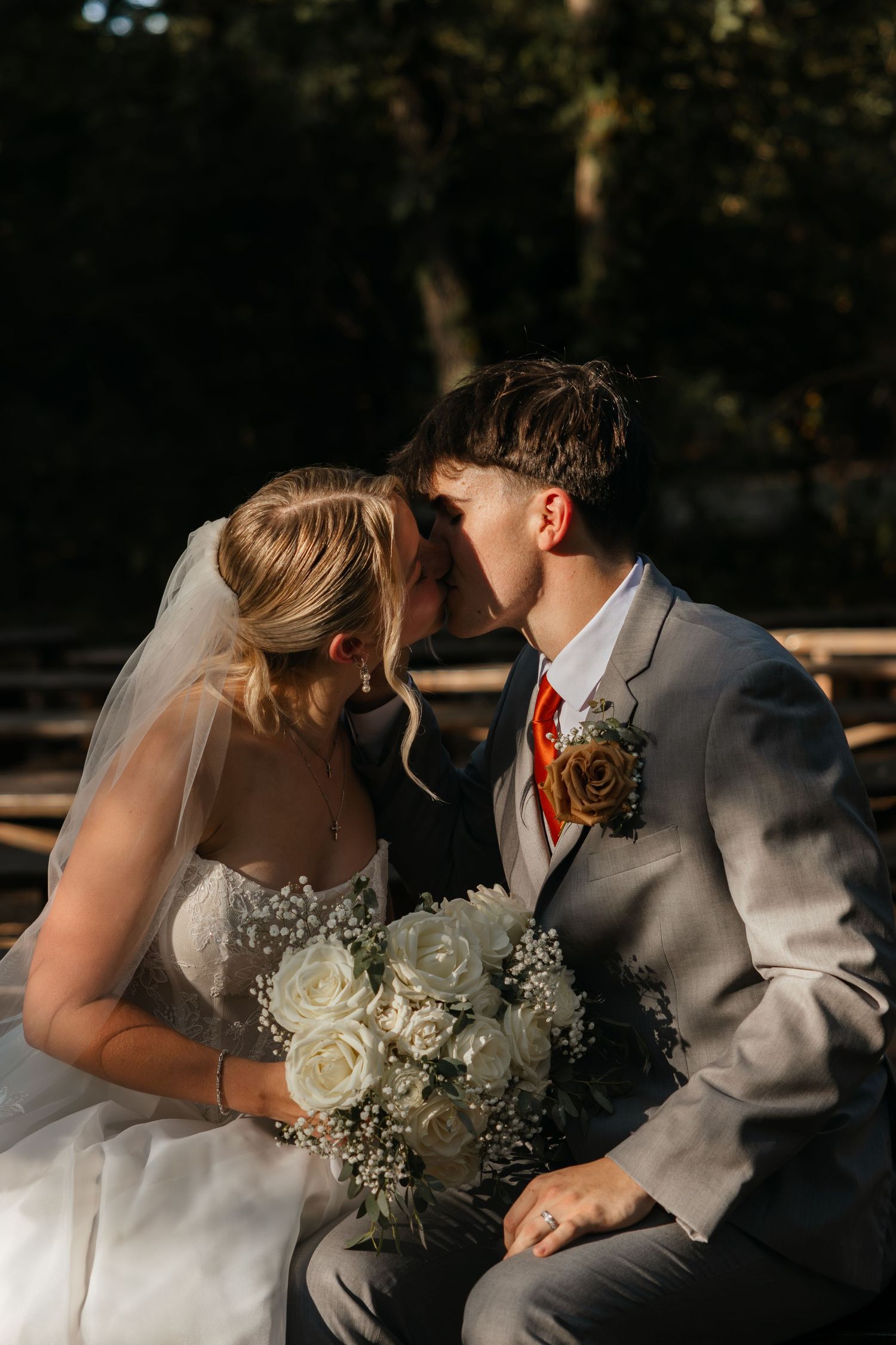 Bride and groom share a tender kiss while seated outdoors, bride holding white rose and baby breath bouquet.