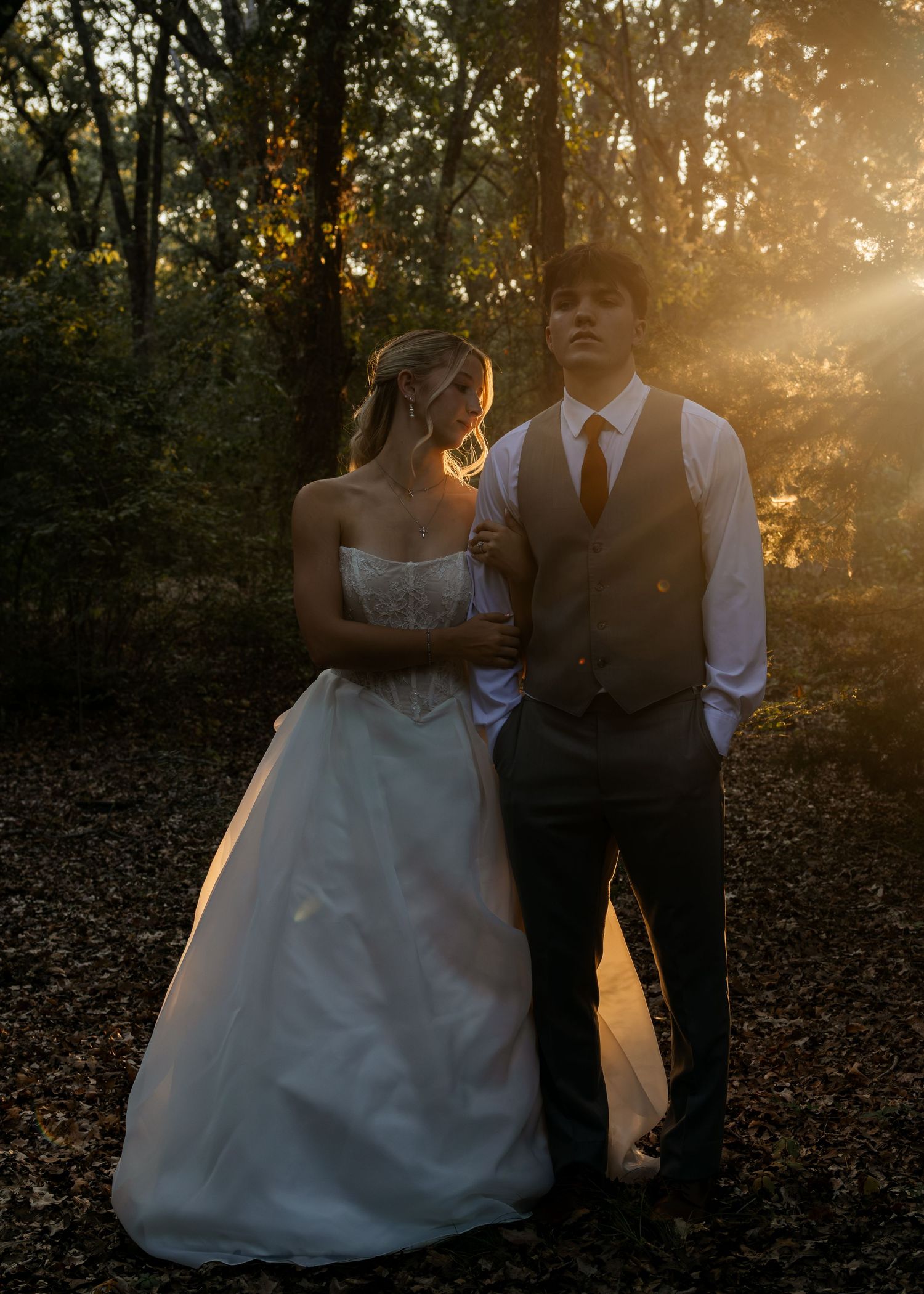 Bride and groom stand together in golden sunset light filtering through trees in romantic forest setting.