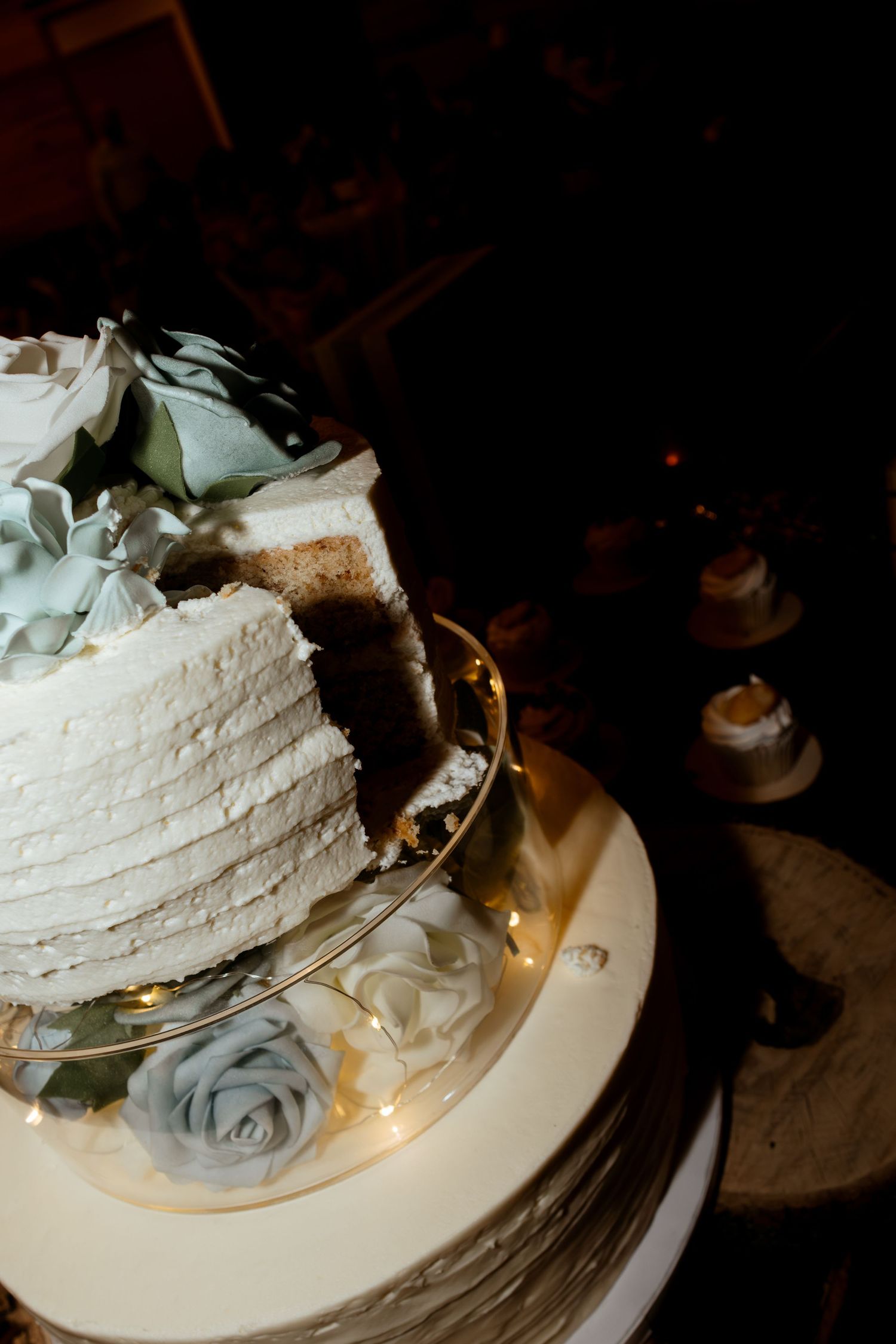 Elegant white wedding cake with blue roses and fairy lights, partially sliced to reveal chocolate layers inside.