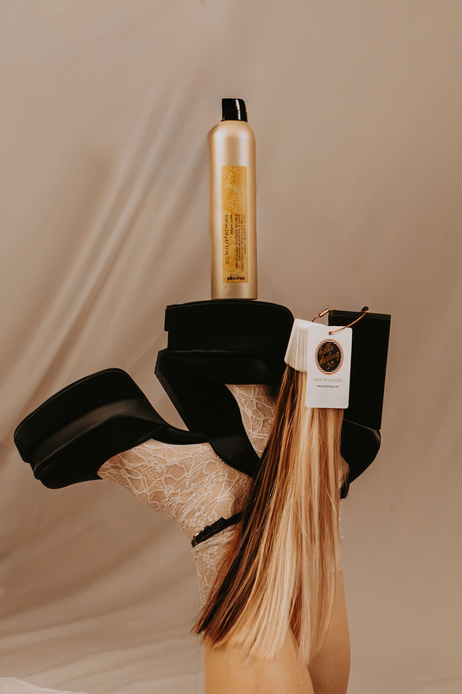 Model balancing gold hairspray on foot wearing black platform heels with lace stockings against beige backdrop.