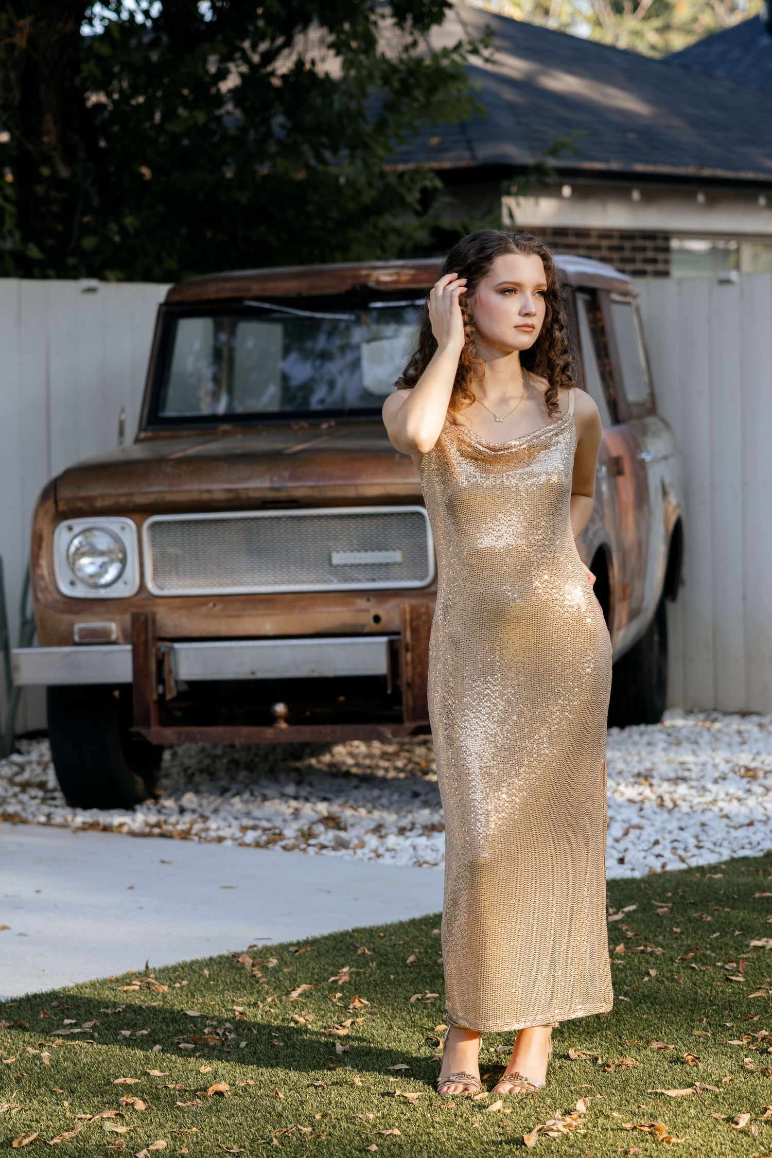 Young woman in gold sequin maxi dress posing outdoors beside a rusty vintage SUV.