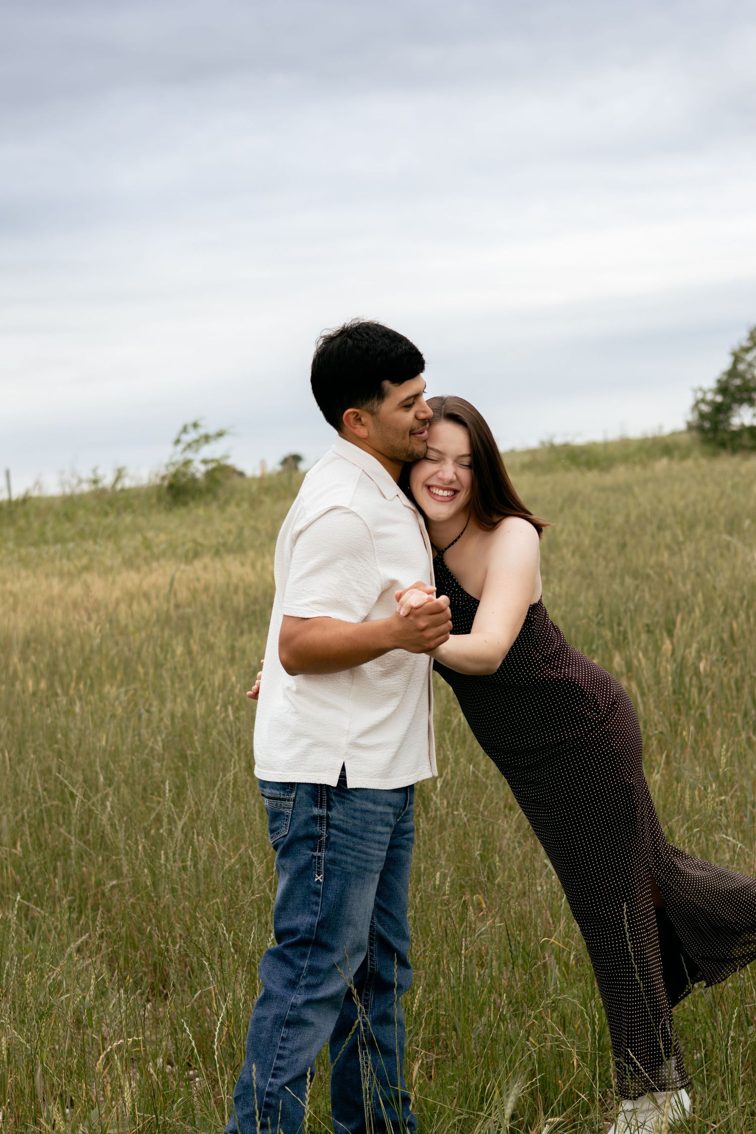 A joyful couple embraces and dances in a lush green field, smiling together under a cloudy sky.