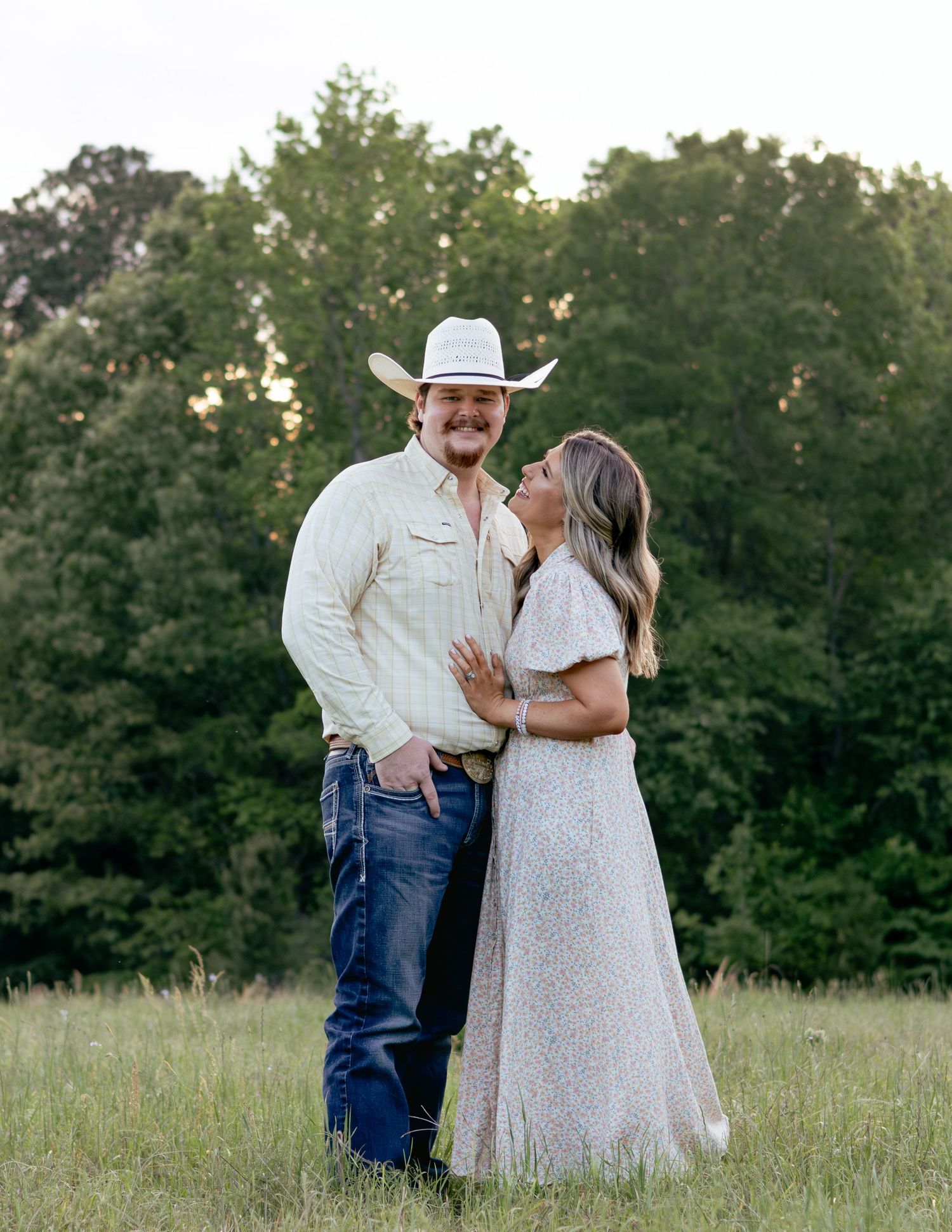 A couple poses romantically in a field at sunset, man wearing a white cowboy hat and cream shirt.