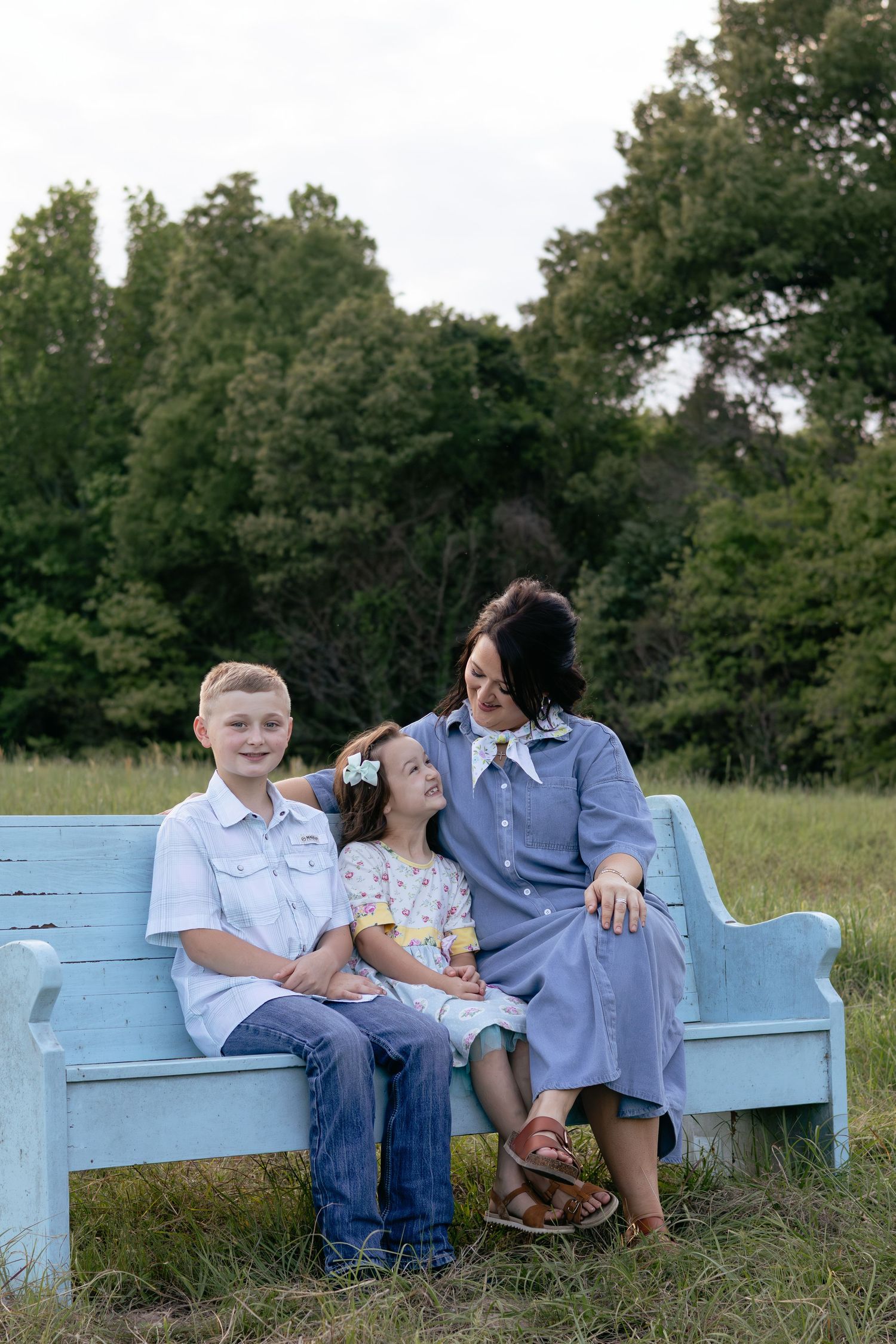 A mother sits on a light blue bench with her two children in an outdoor field setting.