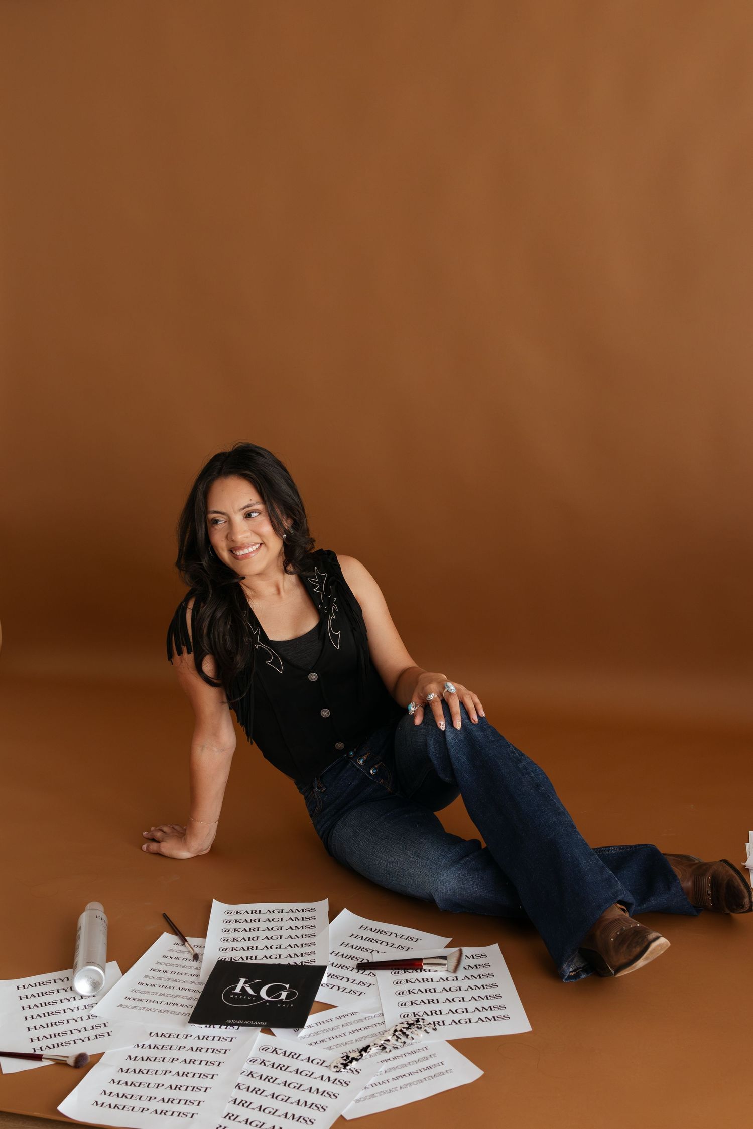 Woman in black top and jeans sits on floor surrounded by KG makeup artist branded papers in studio.