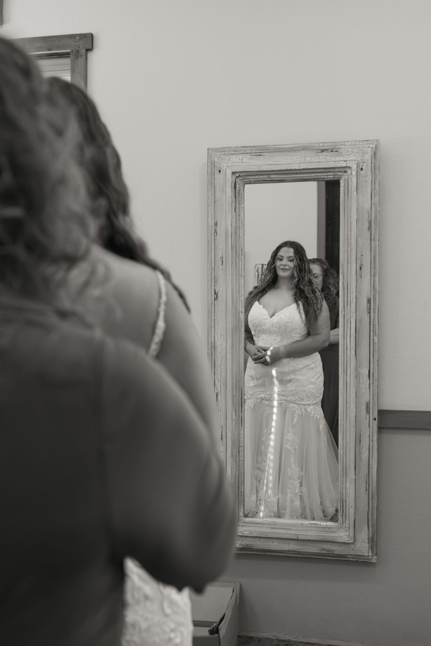 Bride in elegant lace mermaid wedding gown admiring her reflection in a rustic wooden full-length mirror.