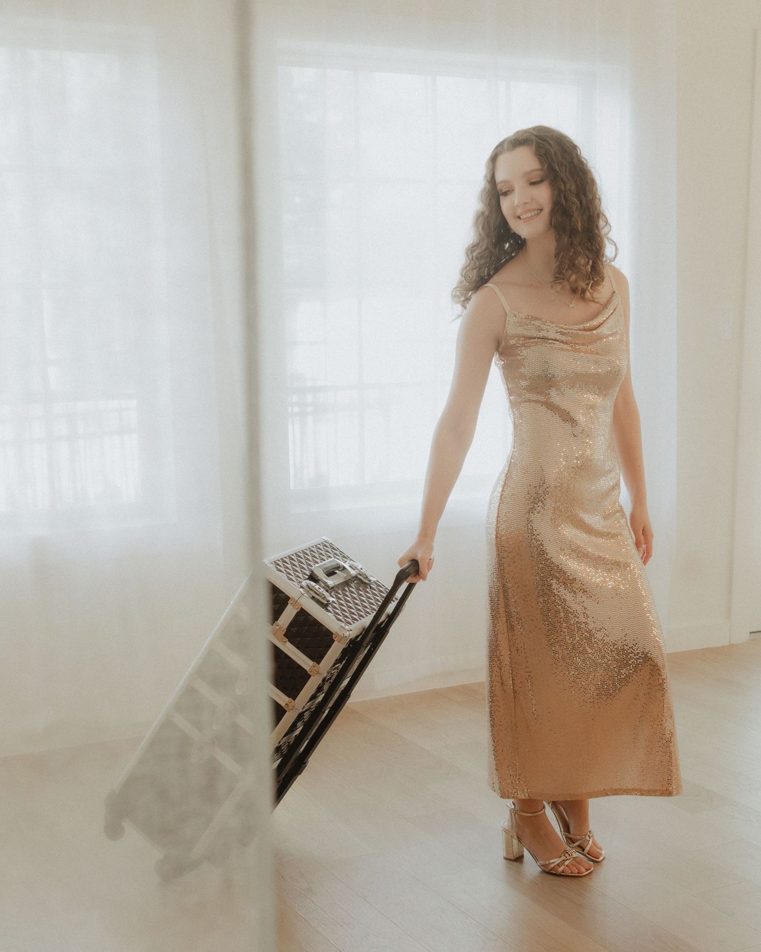 Woman in gold sequin gown smiling while carrying a folding chair in a bright, airy room.