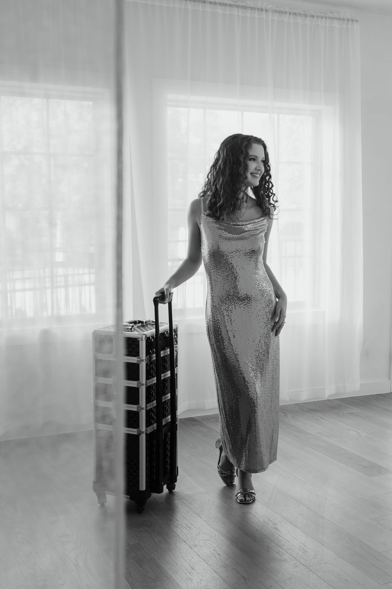 Black and white photo of a woman in sequin gown smiling beside a rolling luggage case.