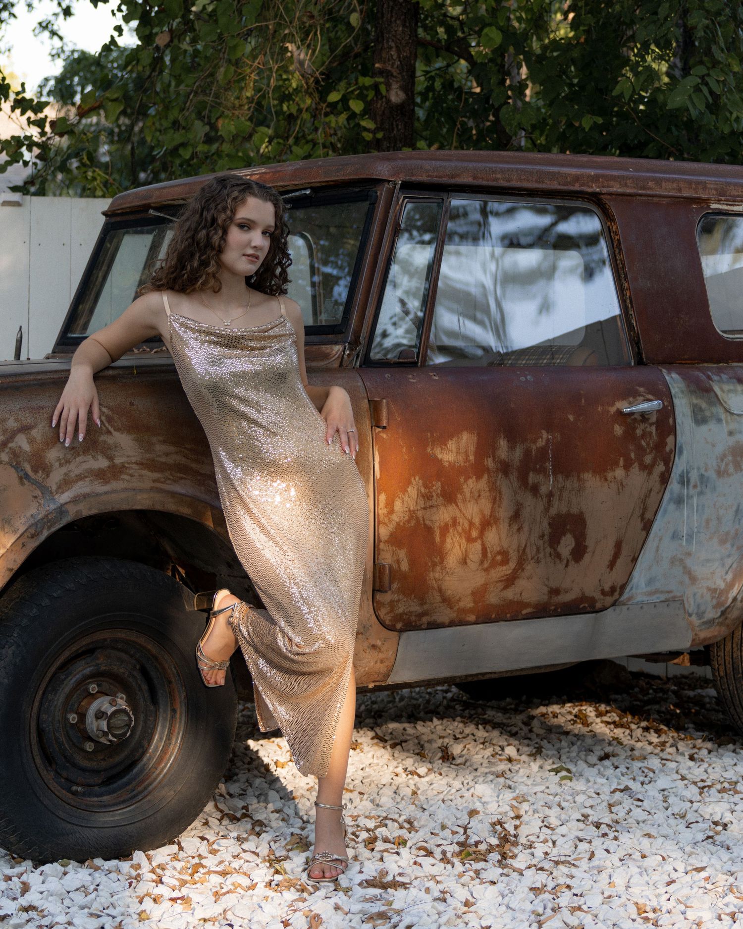 Young woman in gold sequin dress posing beside a rusty vintage car outdoors at golden hour.