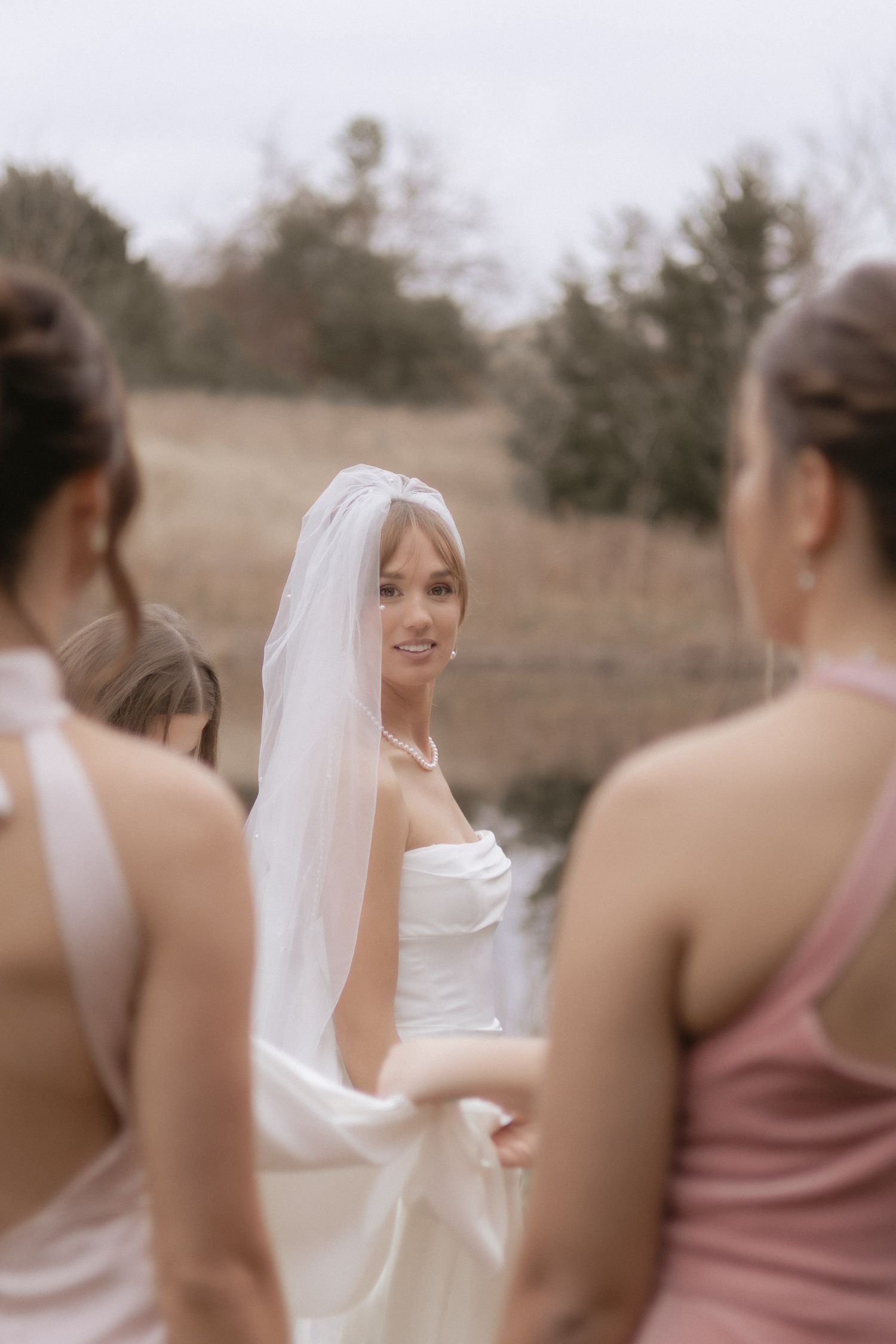 Smiling bride in white strapless gown and veil with bridesmaids in pink dresses outdoors.