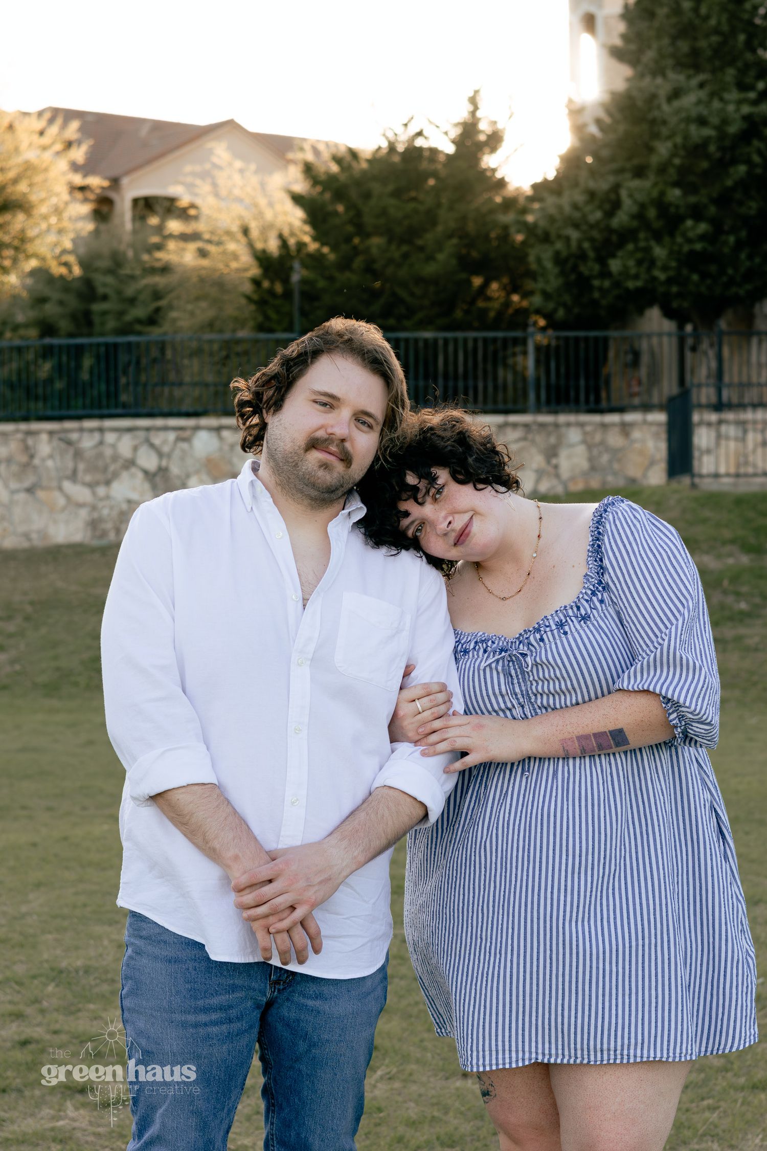 Couple posing outdoors at golden hour, woman in striped dress leaning on man in white shirt, stone wall background.