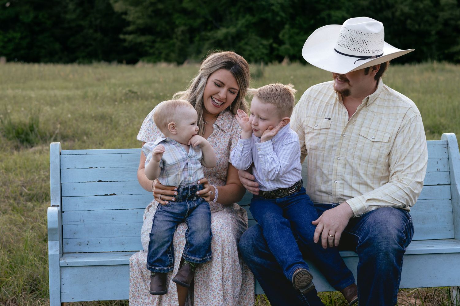 Young family with two small children sits together on a blue bench in a grassy outdoor setting.