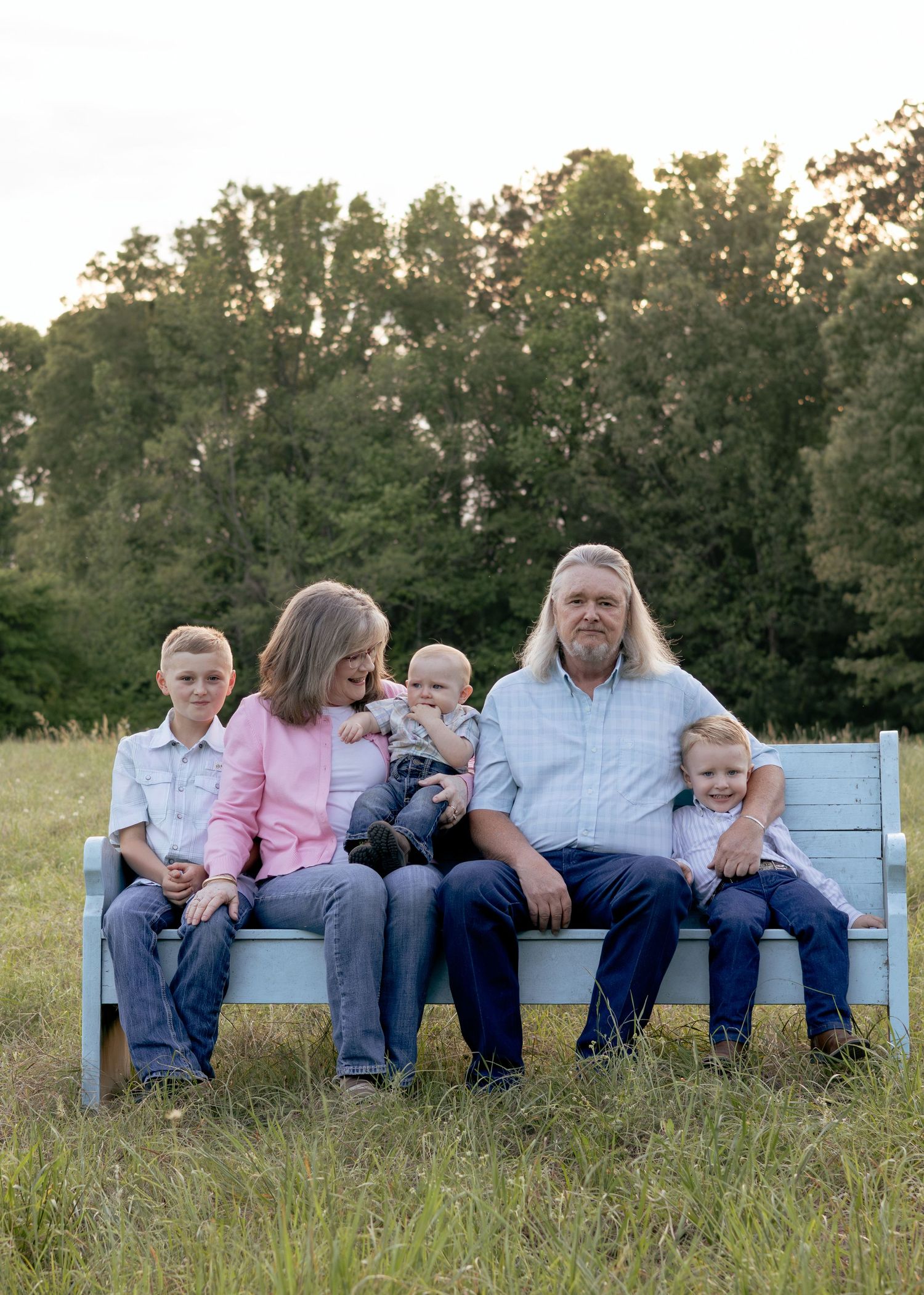 Family of five posing on a light blue bench in a grassy field with trees in the background during golden hour.