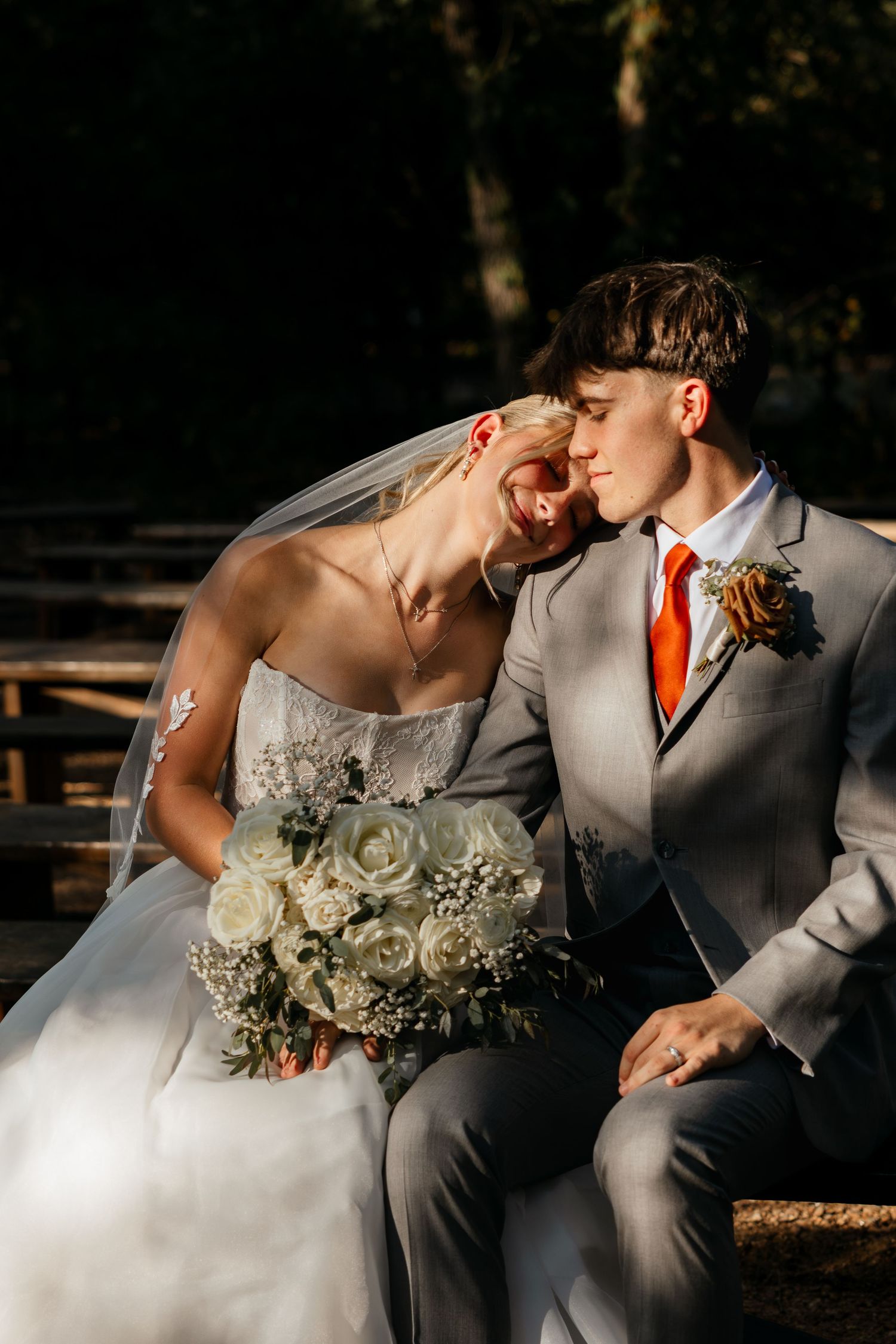 Bride and groom share a tender moment seated outdoors, she holds white roses bouquet, he wears gray suit with orange tie.