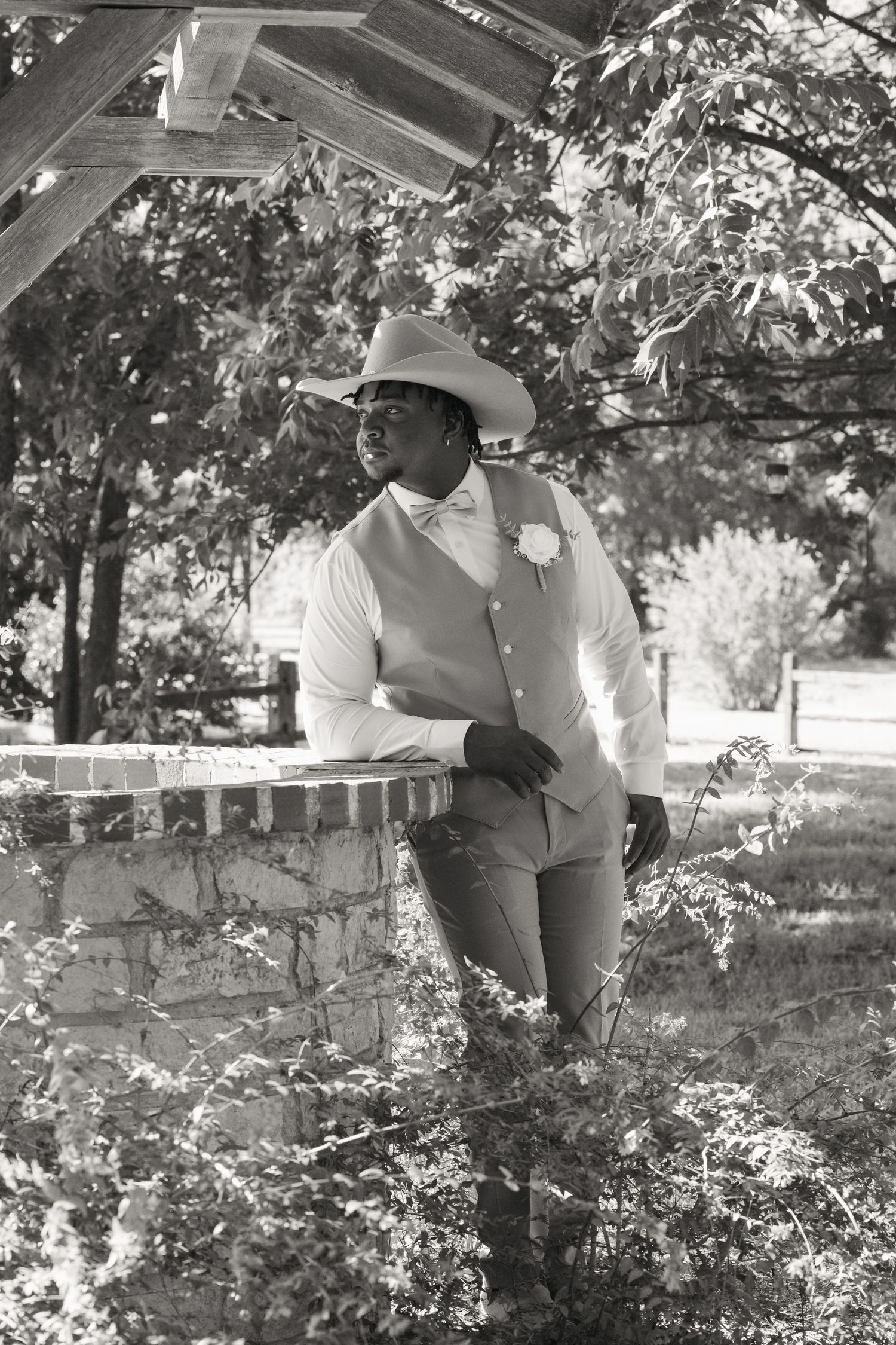 Groom in light blue vest and cowboy hat leaning on brick pillar in black and white outdoor portrait.