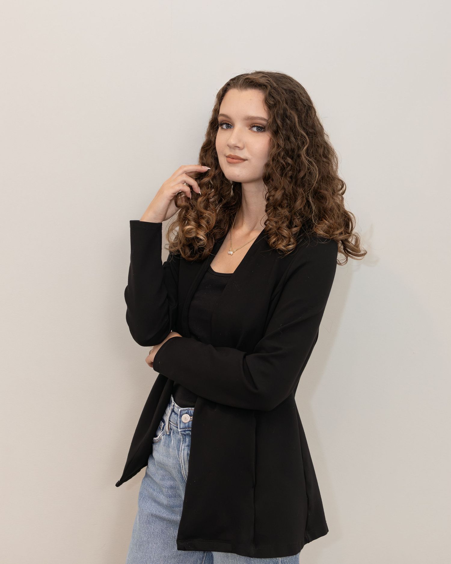 Young woman with curly hair in black blazer and jeans posing thoughtfully against white wall.