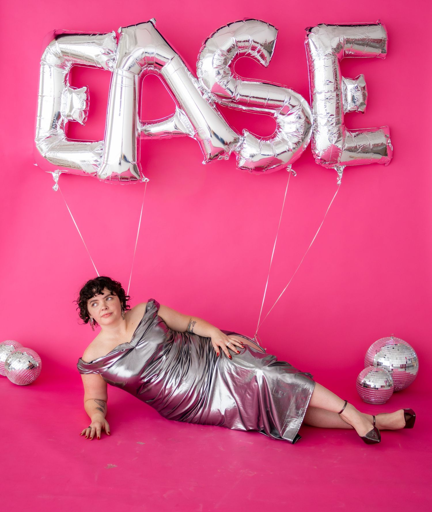 A plus-size woman in a silver metallic dress poses on a pink background with silver EASE letter balloons above her.