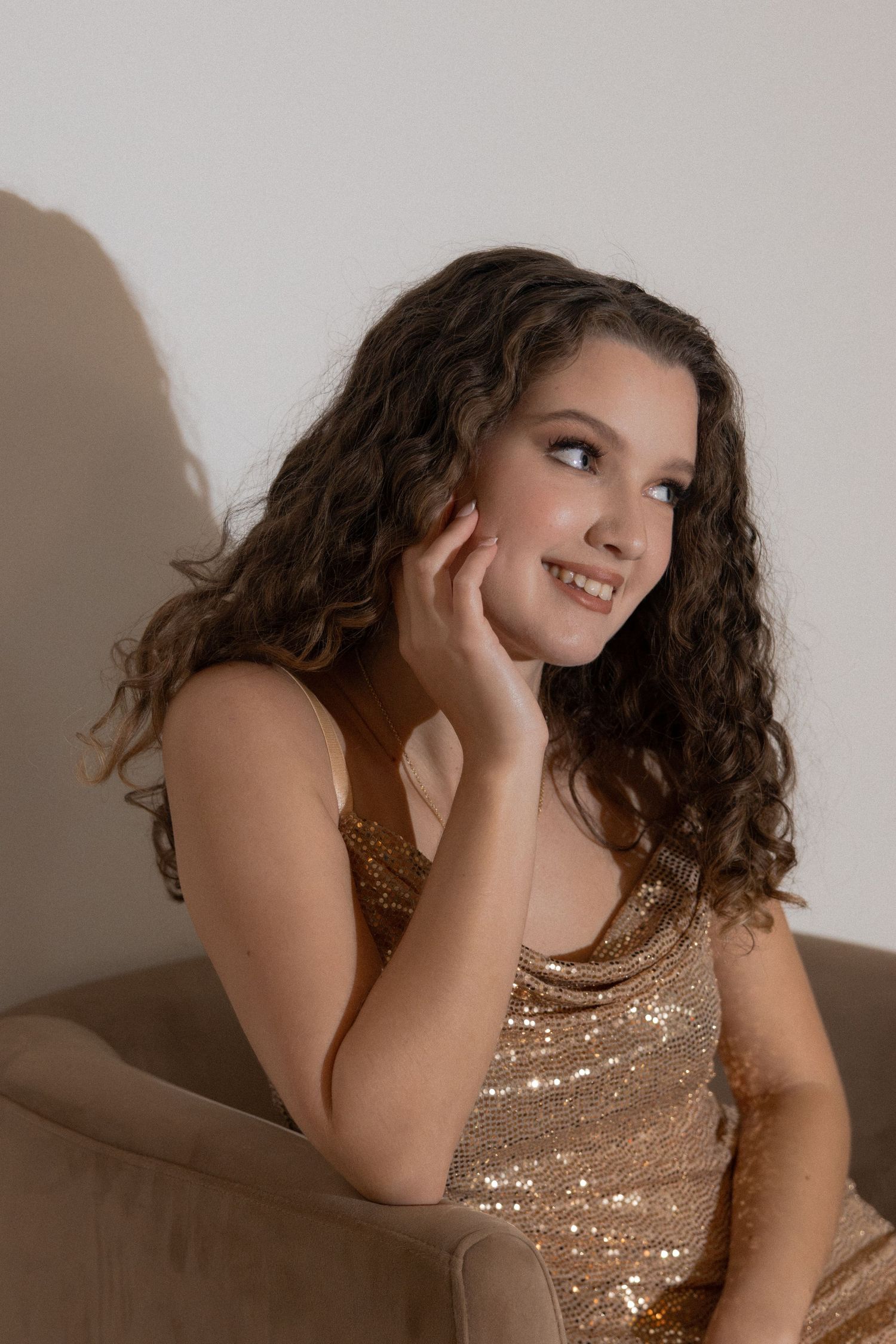 Smiling young woman with curly hair in a gold sequin dress sits relaxed in a chair with hand on cheek.