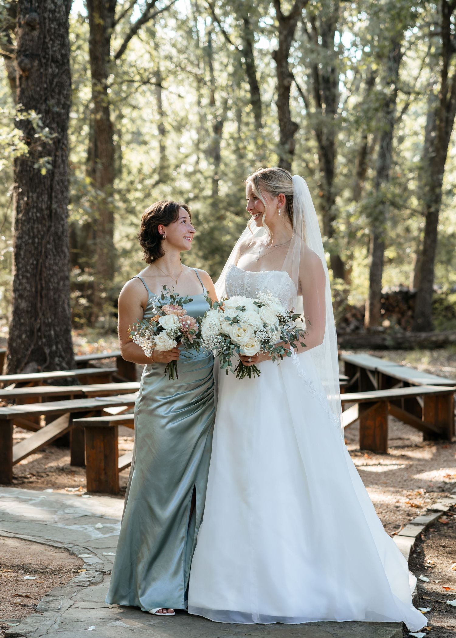 Bride in white gown and bridesmaid in sage green dress holding bouquets at an outdoor woodland wedding venue.