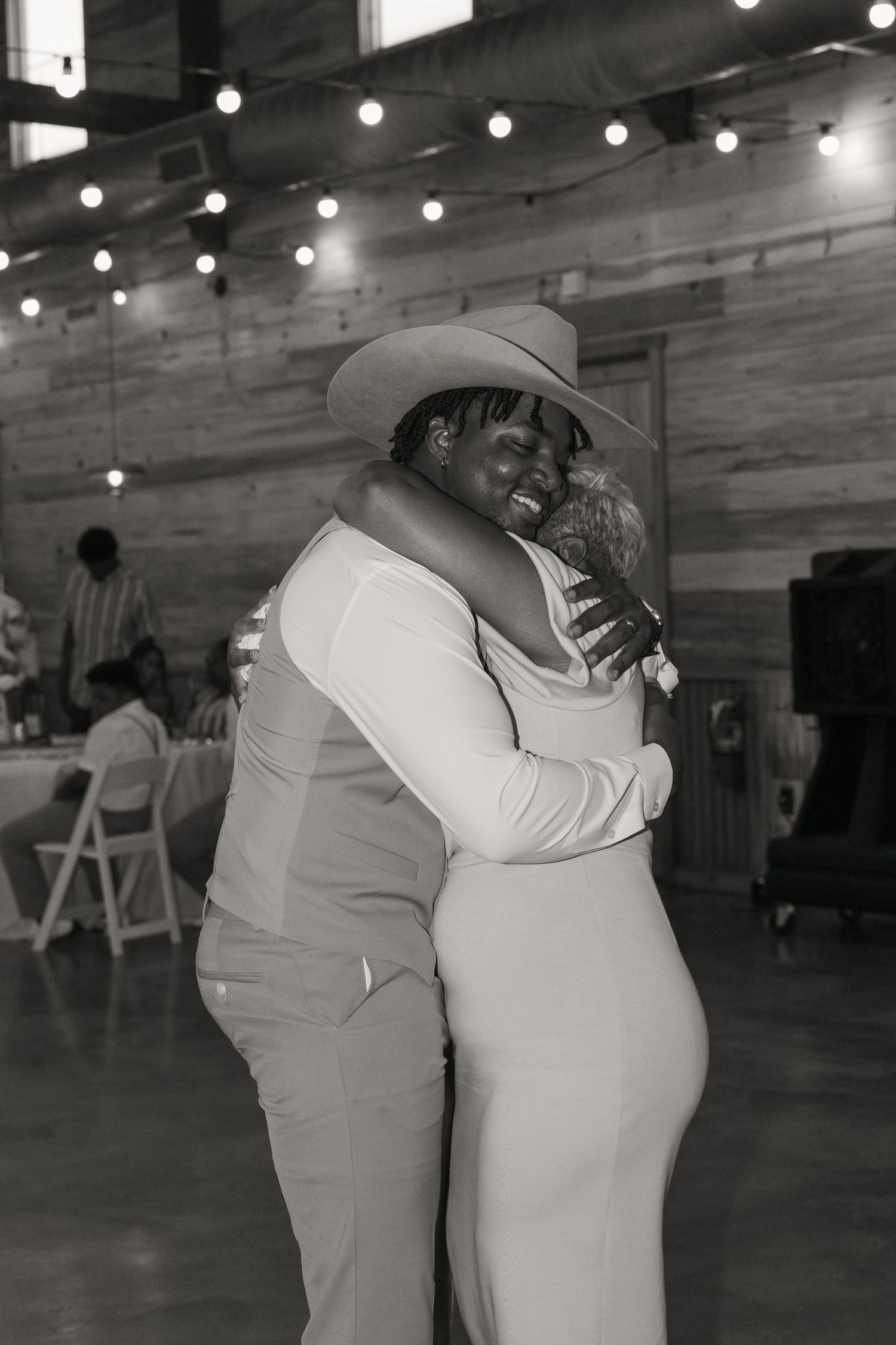 Groom in cowboy hat shares emotional first dance embrace with guest at rustic wedding reception.